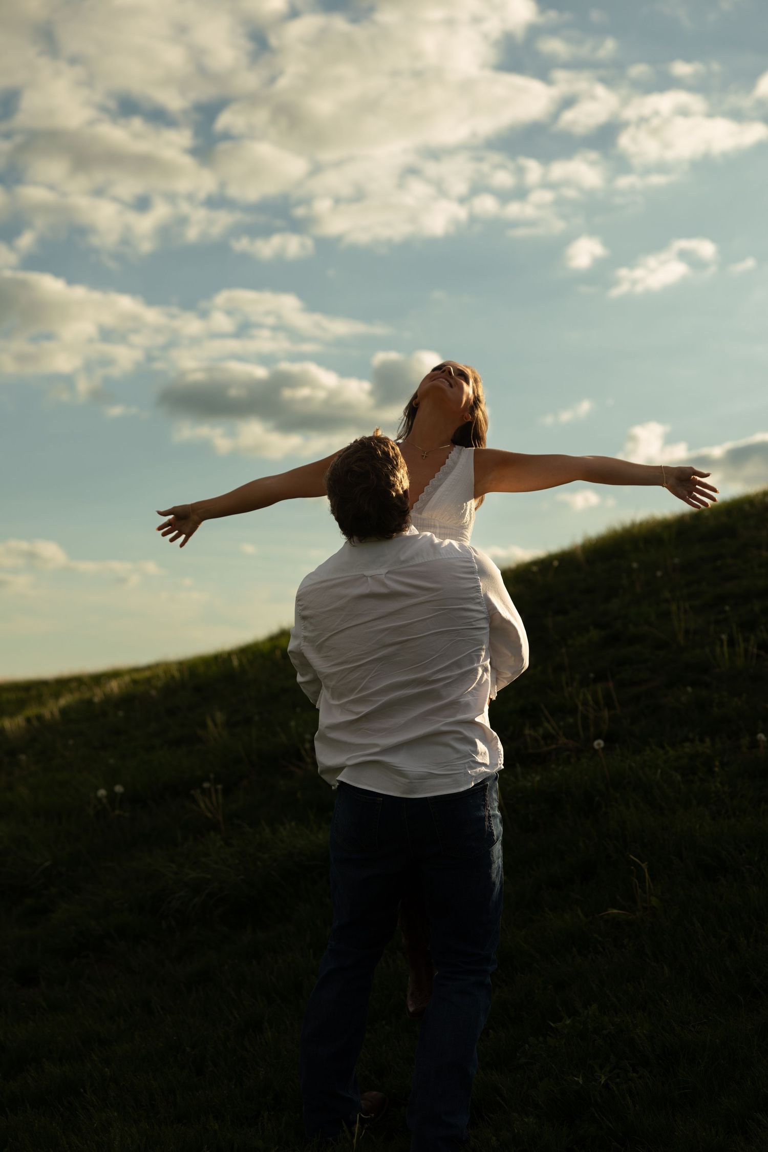 A man lifts a woman with outstretched arms on a grassy hill against a dramatic cloudy sky.