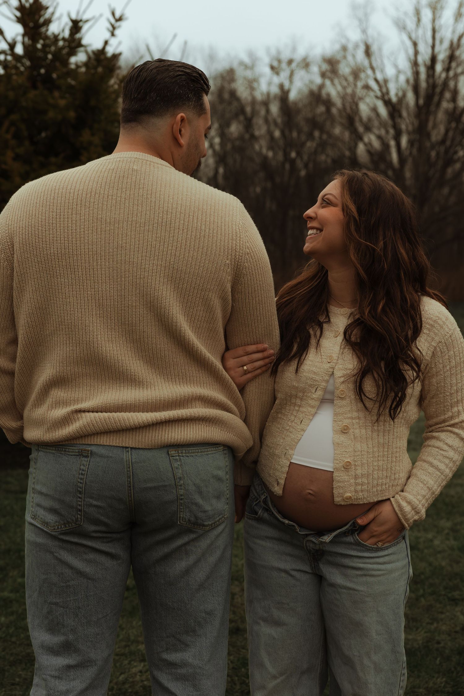 Laughing pregnant woman in cream cardigan showing baby bump during outdoor maternity session.