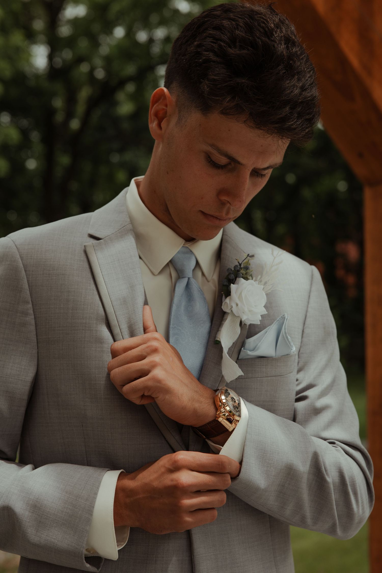 Groom in gray suit adjusting cufflinks with white boutonniere and light blue tie outdoors.