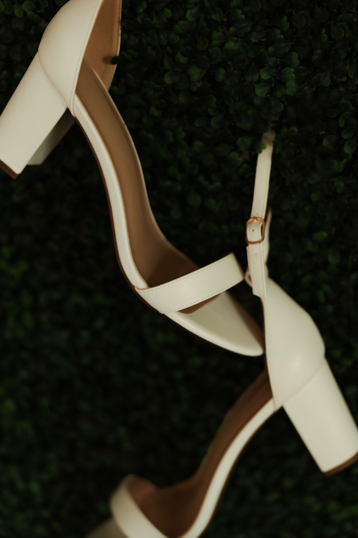 White leather block heel bridal sandals with ankle strap displayed against lush green hedge.