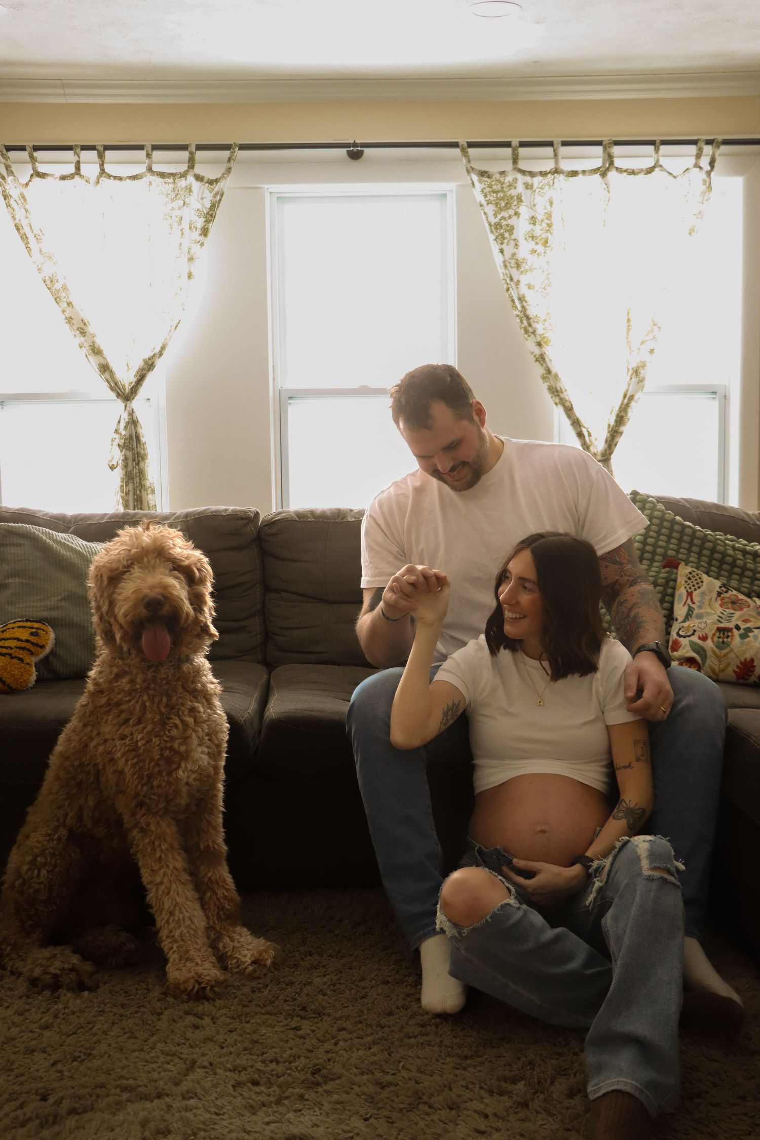 Pregnant woman sits on floor with tattooed partner and fluffy goldendoodle dog in cozy living room.