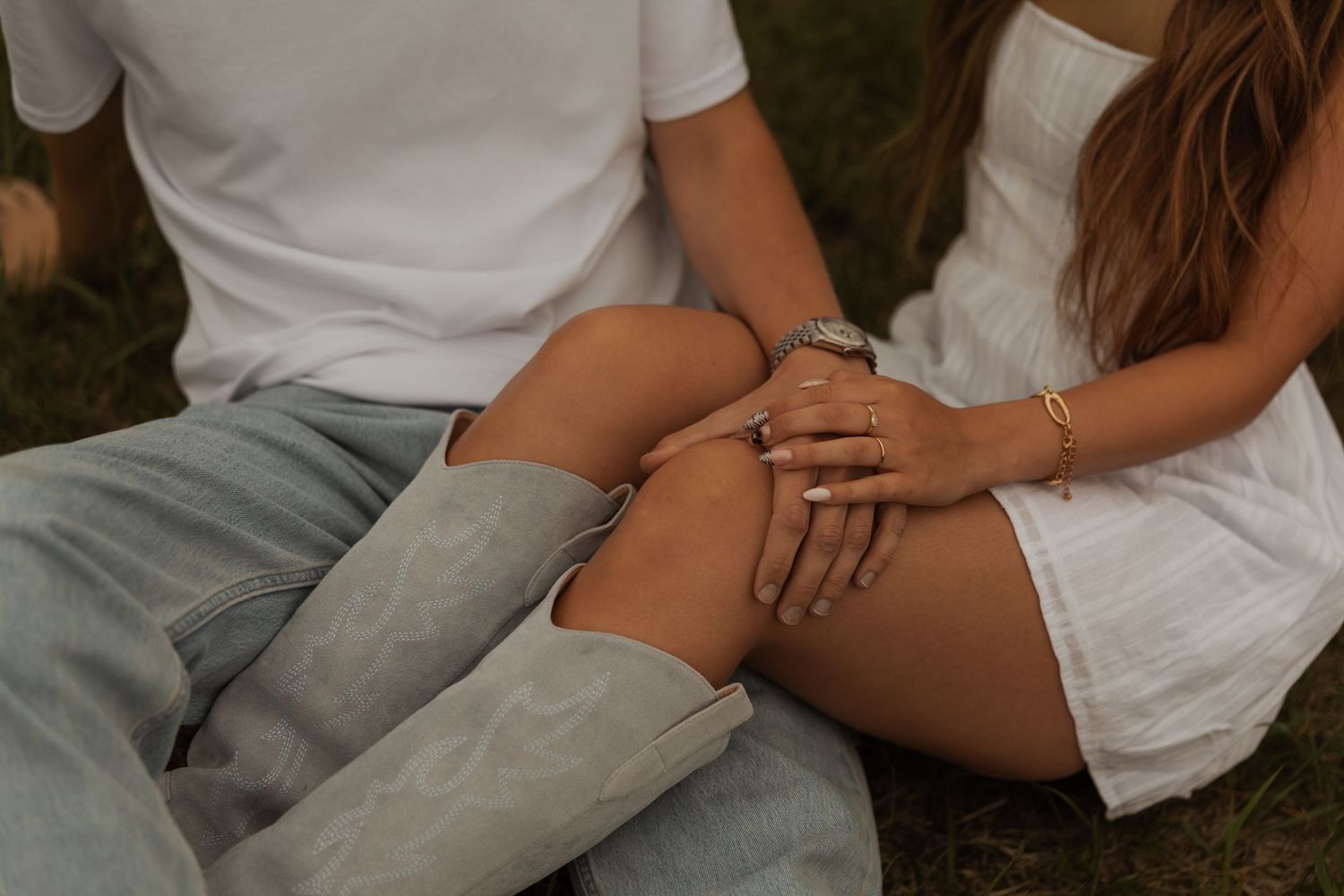 Close-up of a couple sitting in grass, hands intertwined, showcasing an engagement ring and gold bracelet.