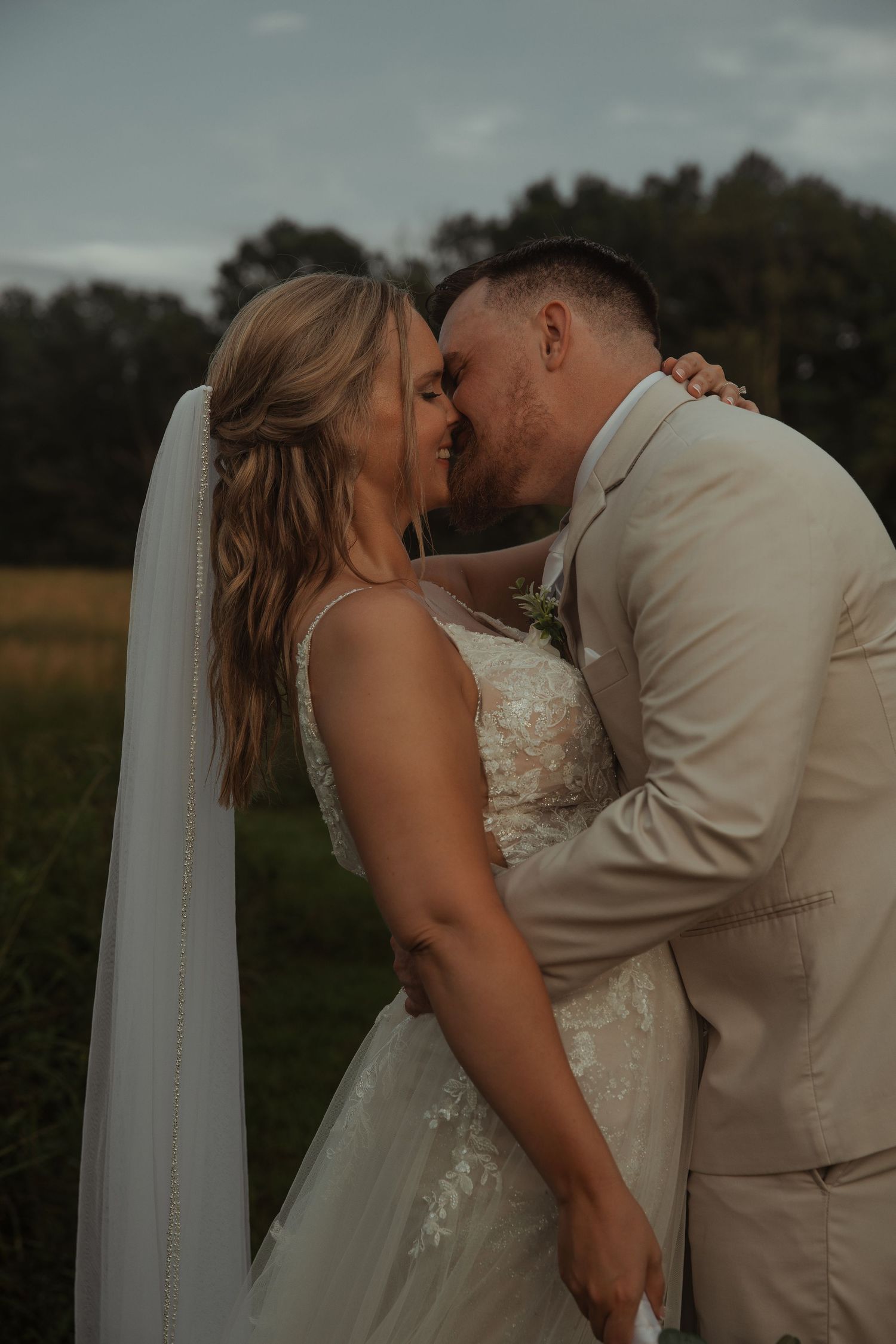 A bride and groom share a romantic kiss outdoors at dusk, with the bride wearing a lace gown and veil.