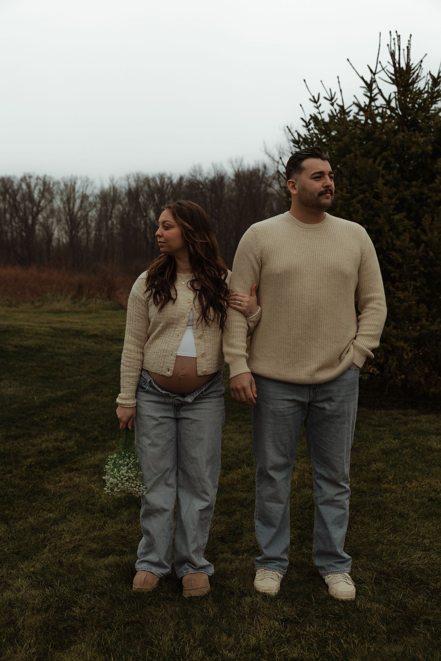 Pregnant couple standing together outdoors in matching beige sweaters and jeans holding flowers.