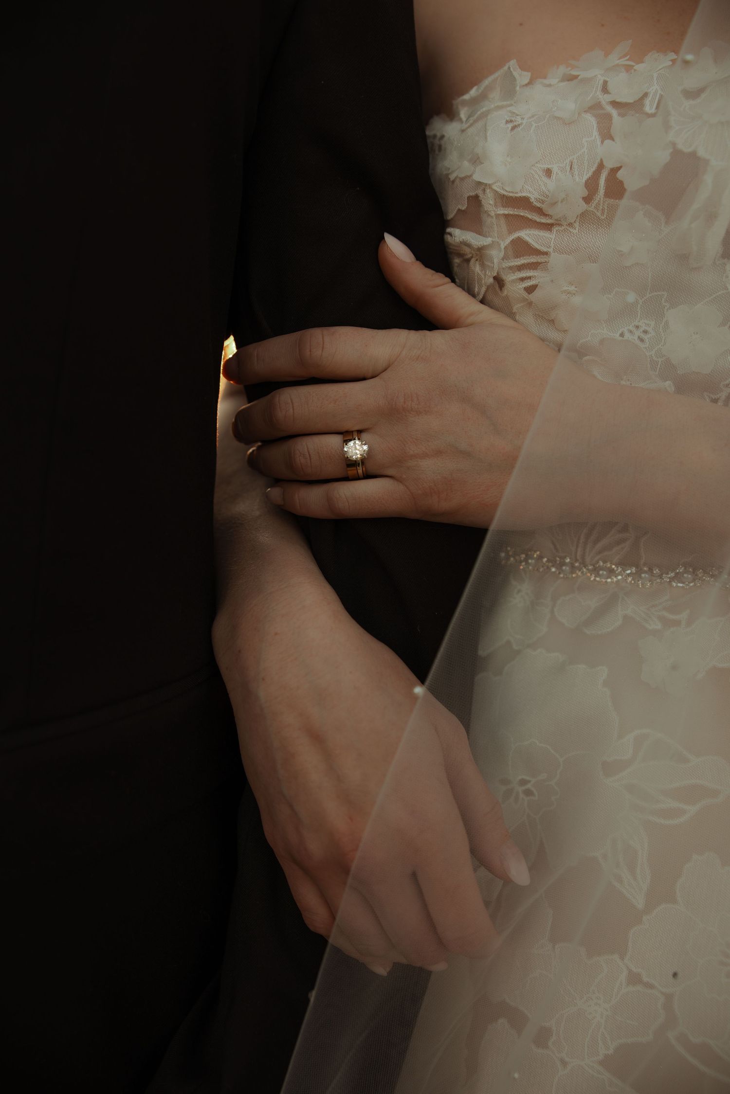 Close-up of bride's hand with a diamond solitaire ring resting on groom's dark suit sleeve.