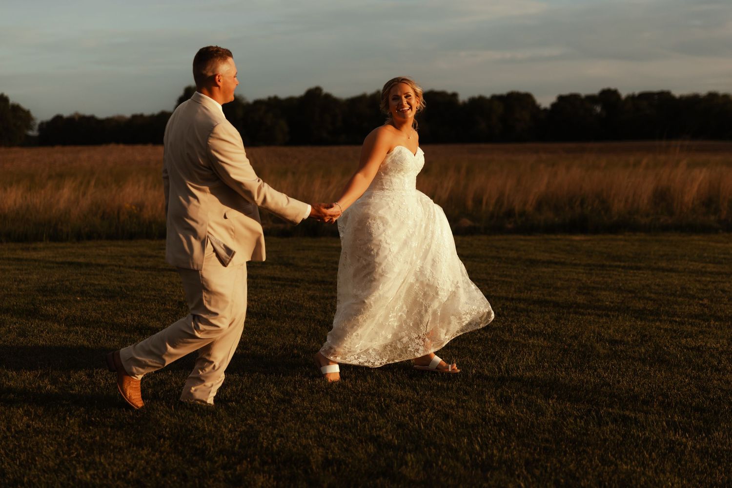 Bride and groom holding hands walking through golden field at sunset during wedding photo session.