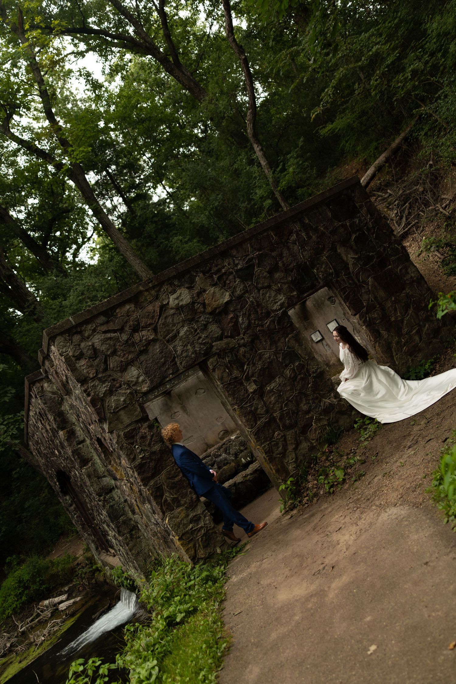 Bride in white gown and groom in blue suit pose near a rustic stone building surrounded by lush green trees.