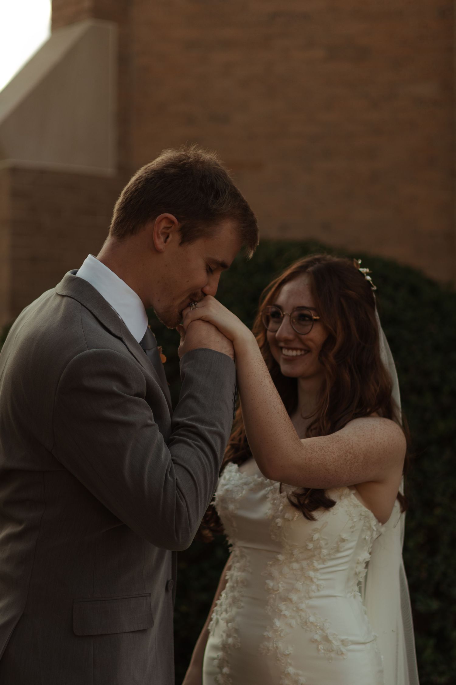 Groom in a gray suit kisses the bride's hand as she smiles warmly outside a brick church.