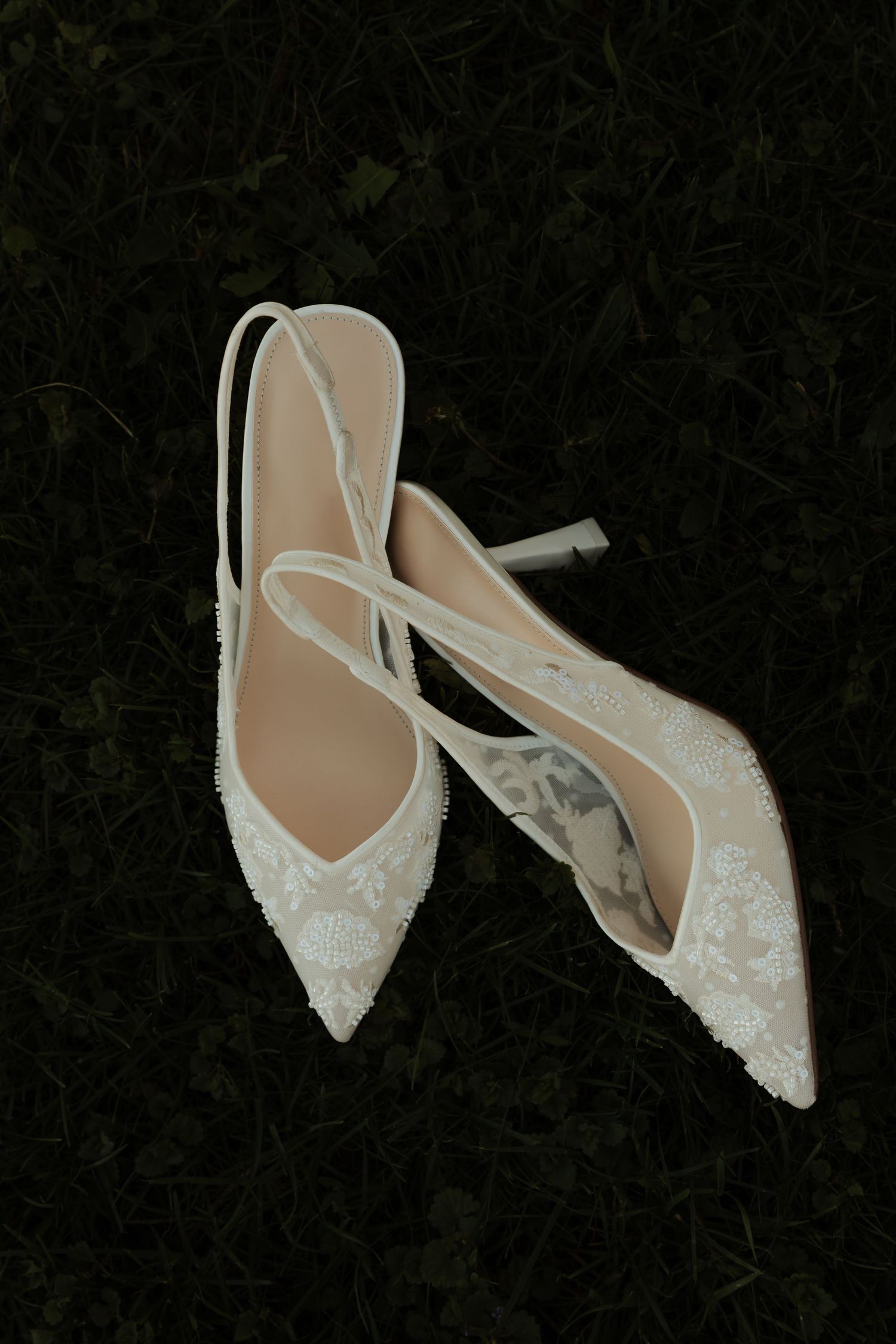 White lace pointed-toe slingback bridal heels resting on dark green grass.