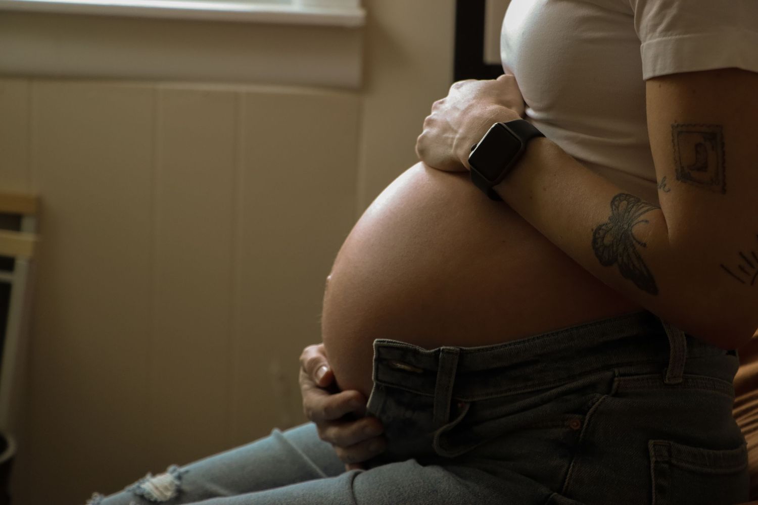Close-up of pregnant woman's bare belly with tattoos and smartwatch in ripped jeans.