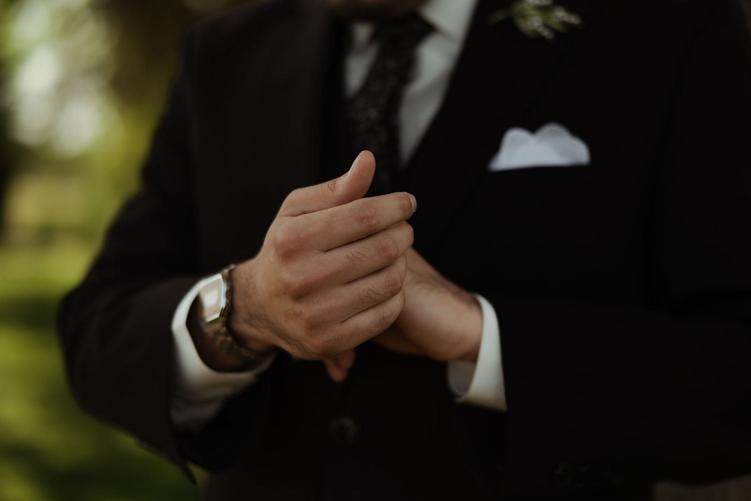 Groom in black suit clasping hands, wearing a watch and white pocket square at a wedding.
