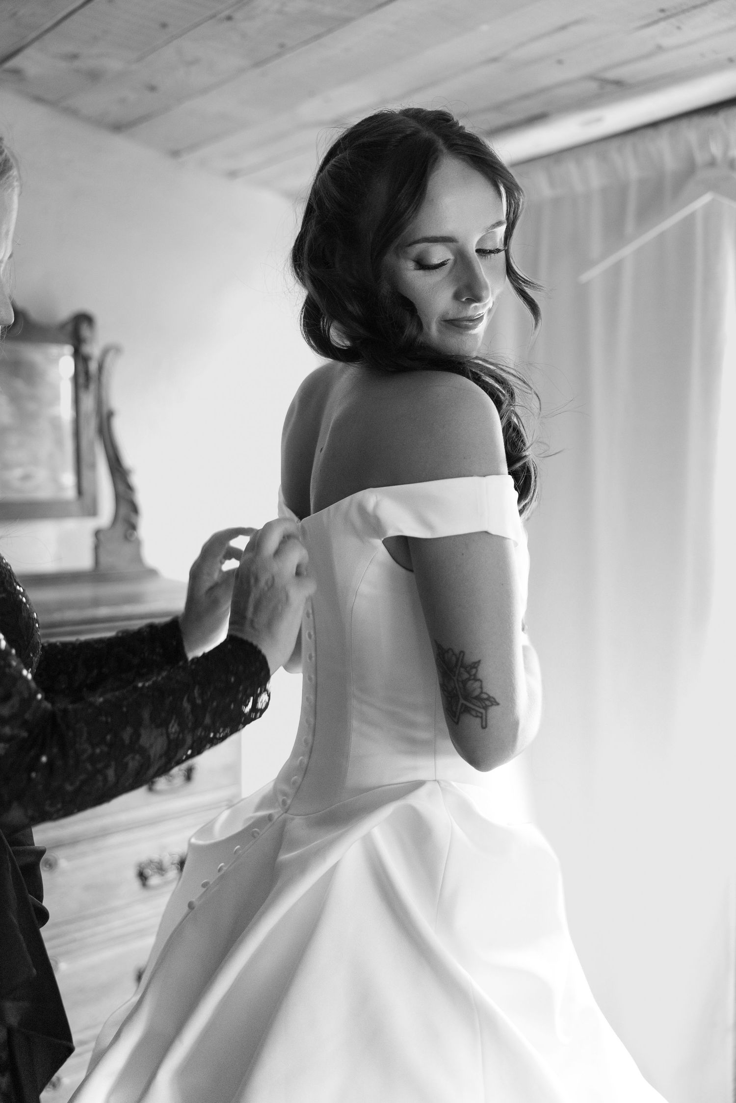 Black and white photo of a bride in an off-shoulder wedding gown being buttoned up, showing her floral tattoo.