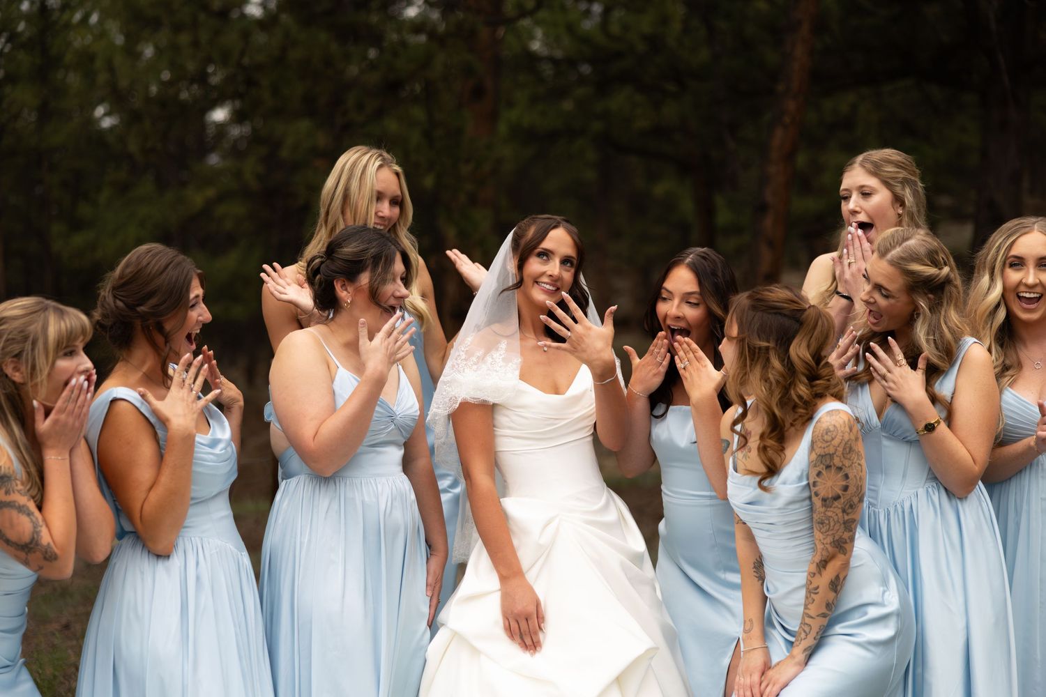 Bride in white gown showing ring to excited bridesmaids in light blue dresses outdoors.