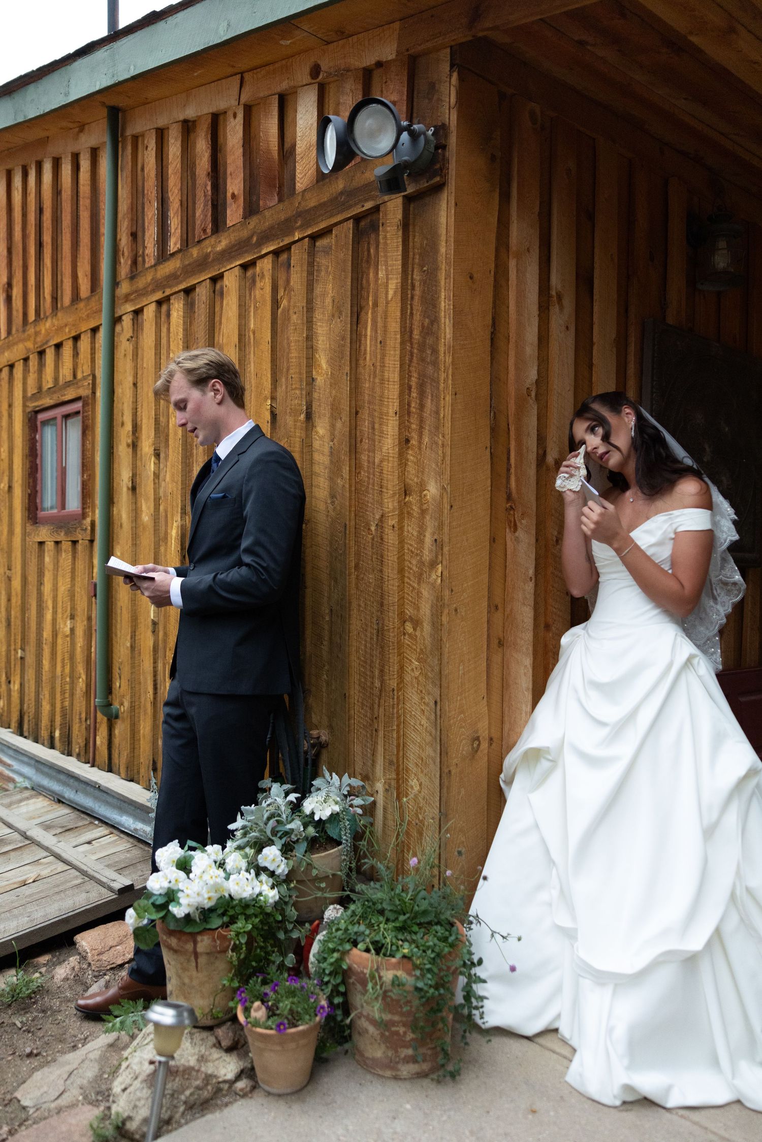 Bride in white ball gown crying while groom reads vows outside rustic wooden barn with potted flowers.