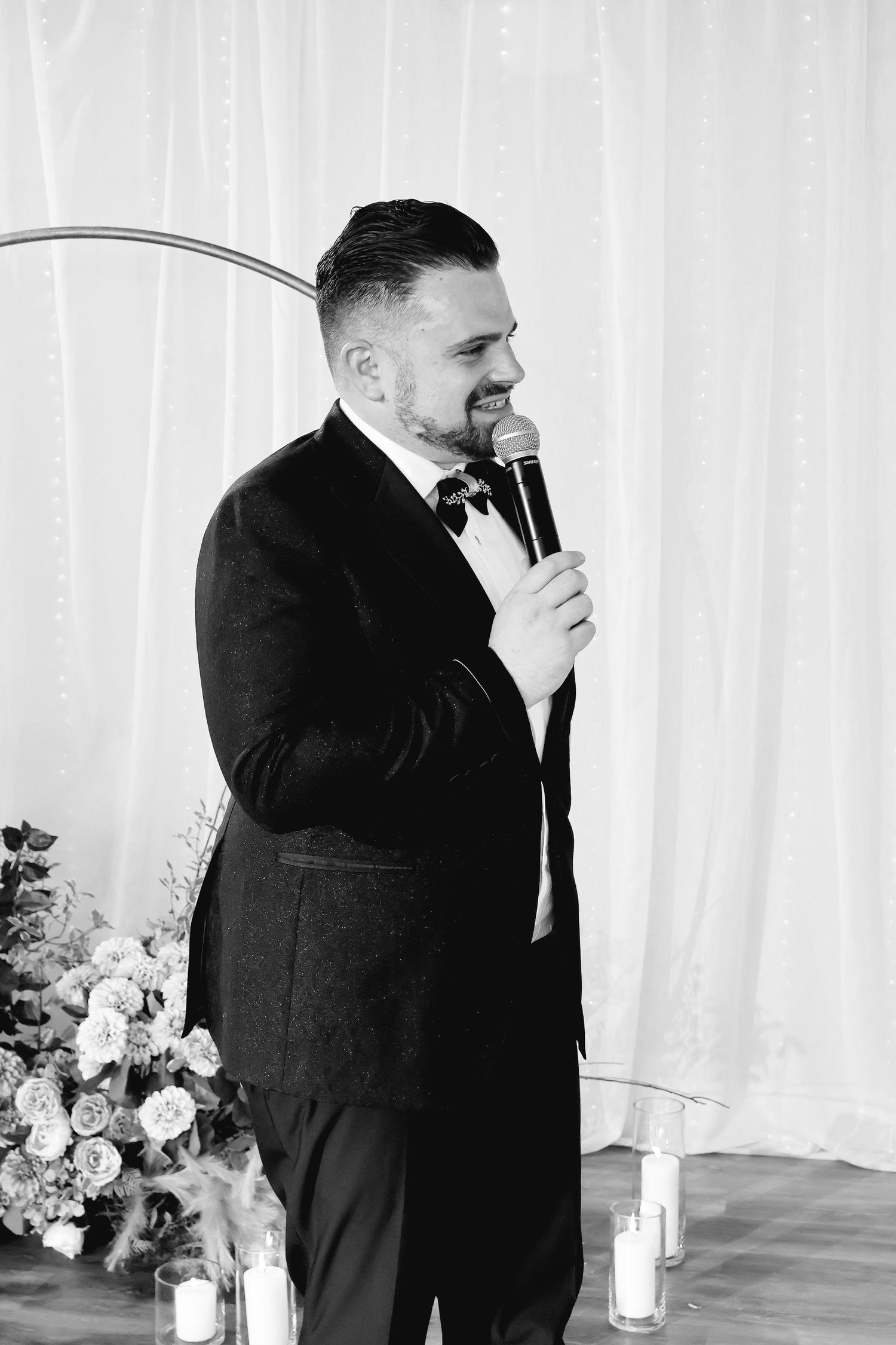 Black and white photo of a groom in a tuxedo speaking into a microphone at a wedding ceremony.