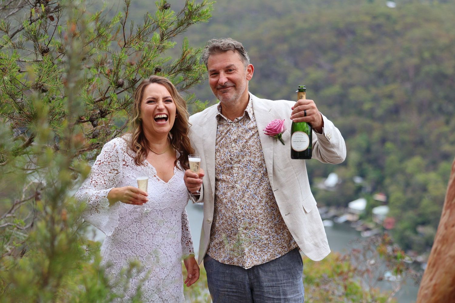 Smiling couple celebrating with champagne on scenic hilltop overlook, bride in lace dress and groom in floral shirt.