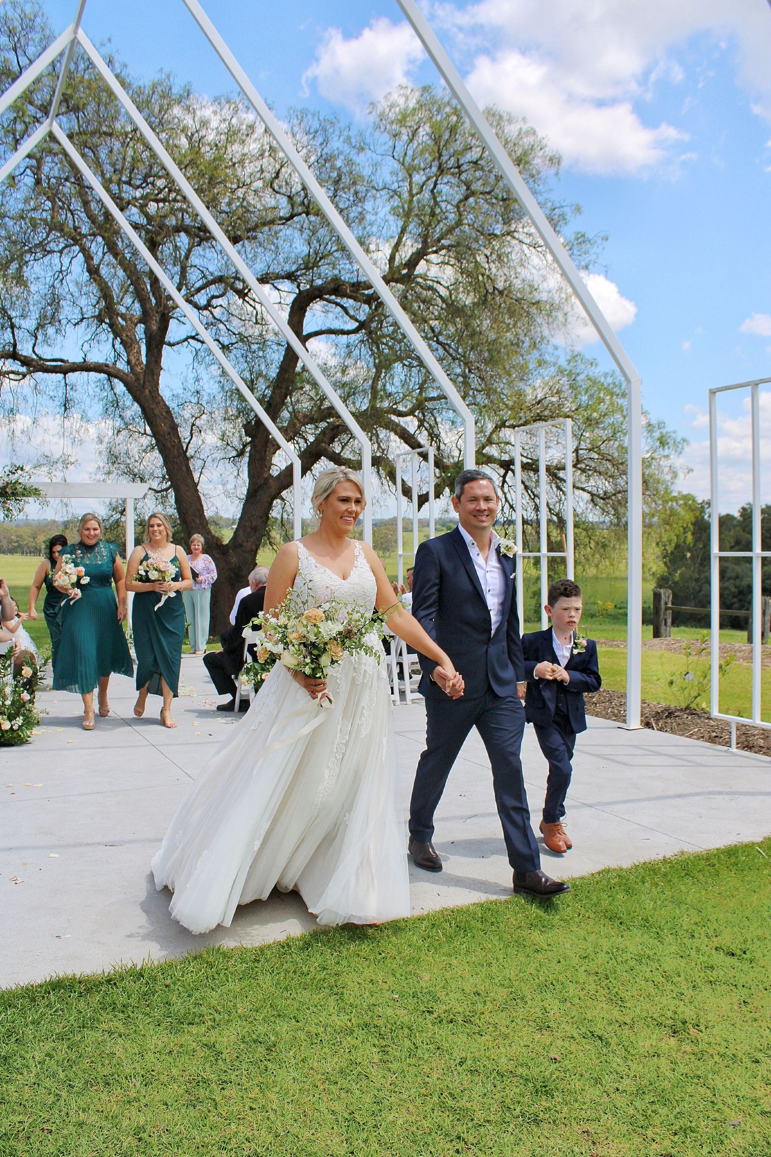 Newlyweds walking down aisle with son after outdoor ceremony, bridesmaids in green dresses in background.