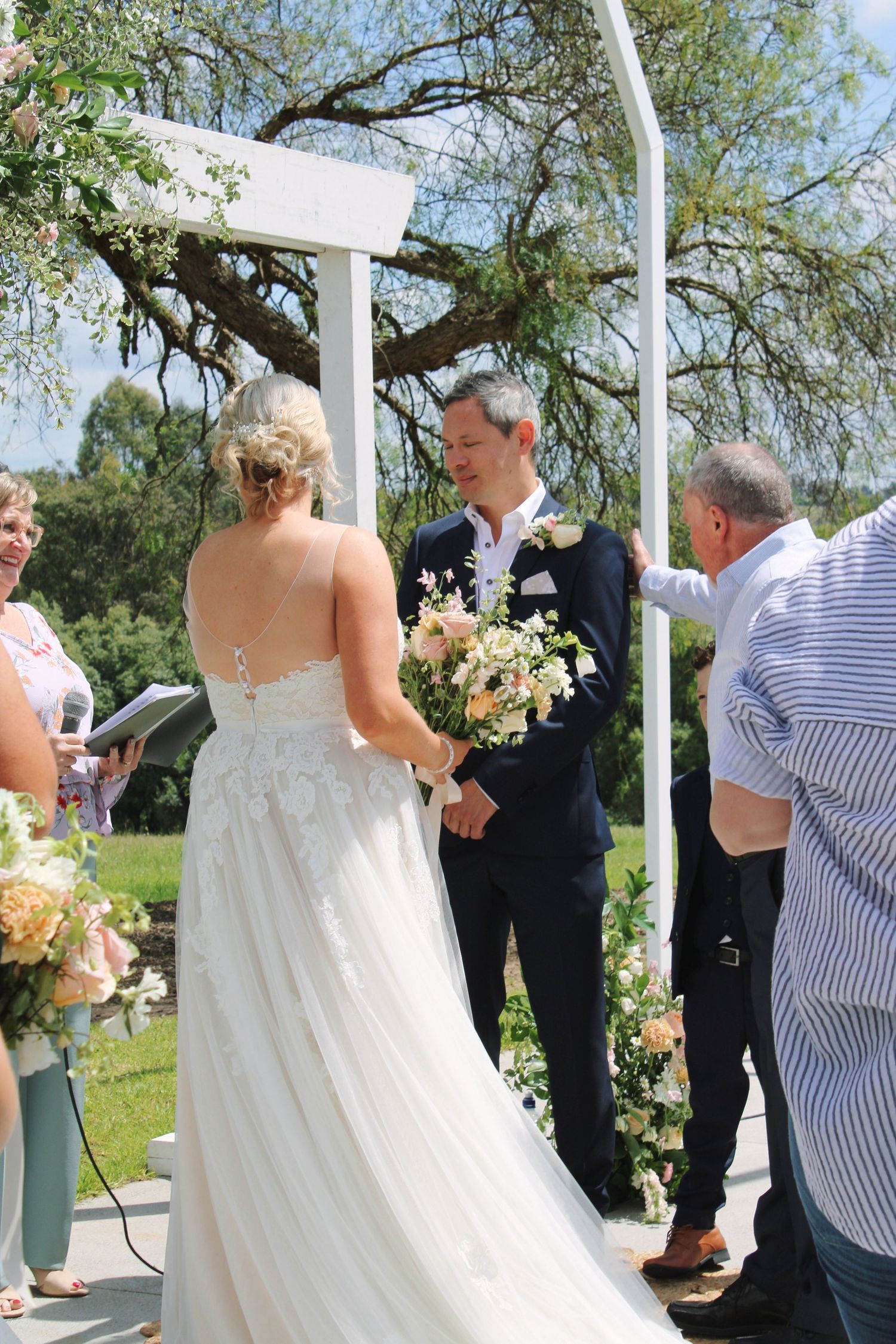 Bride and groom exchange vows at a white floral arch during an outdoor garden wedding ceremony.