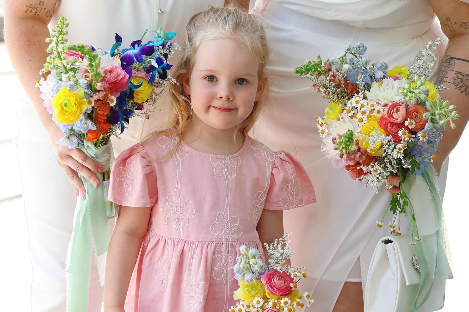 Adorable blonde flower girl in pink dress stands between two brides holding colorful wedding bouquets.