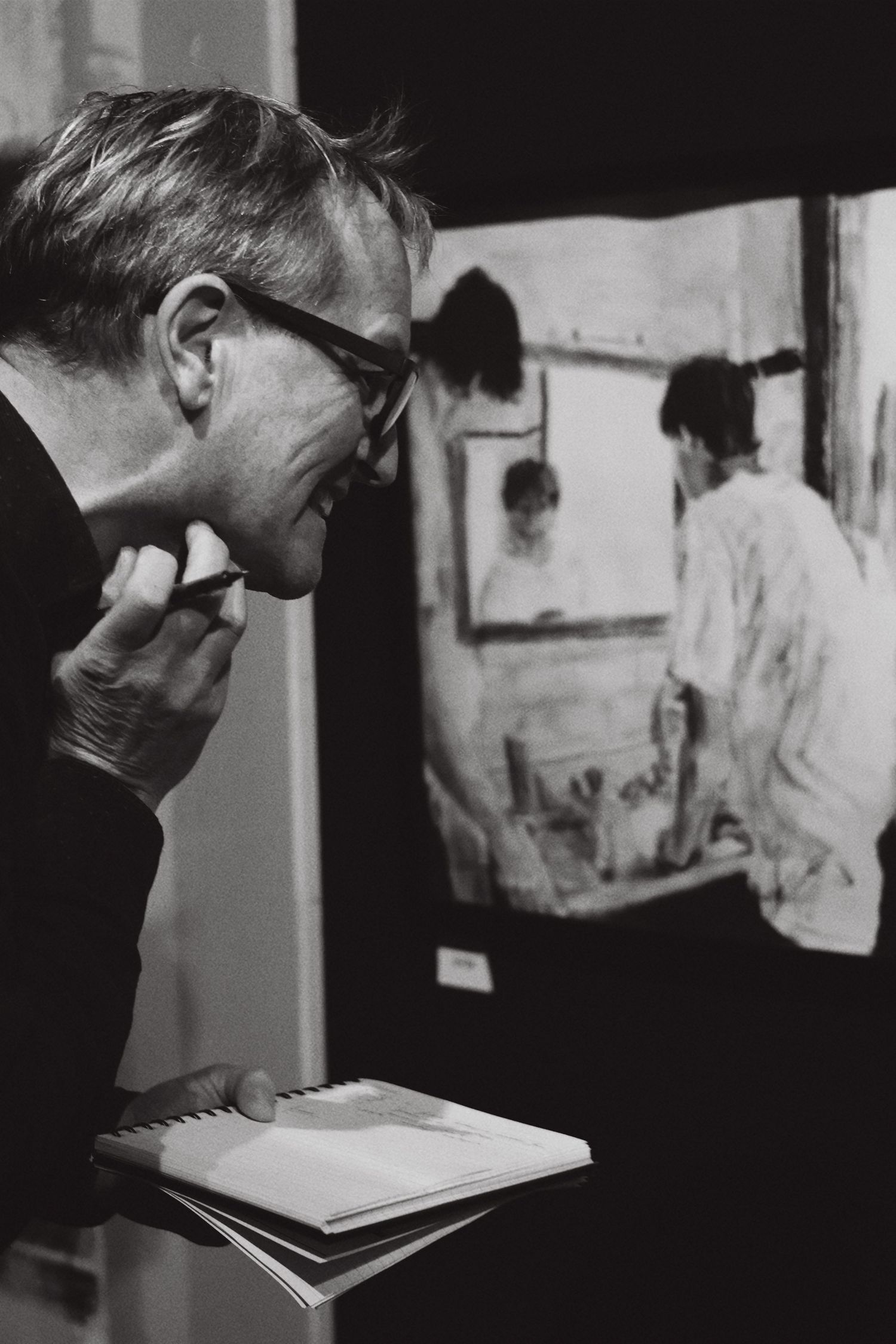 Black and white photo of man with glasses studying artwork while holding notebook at gallery.