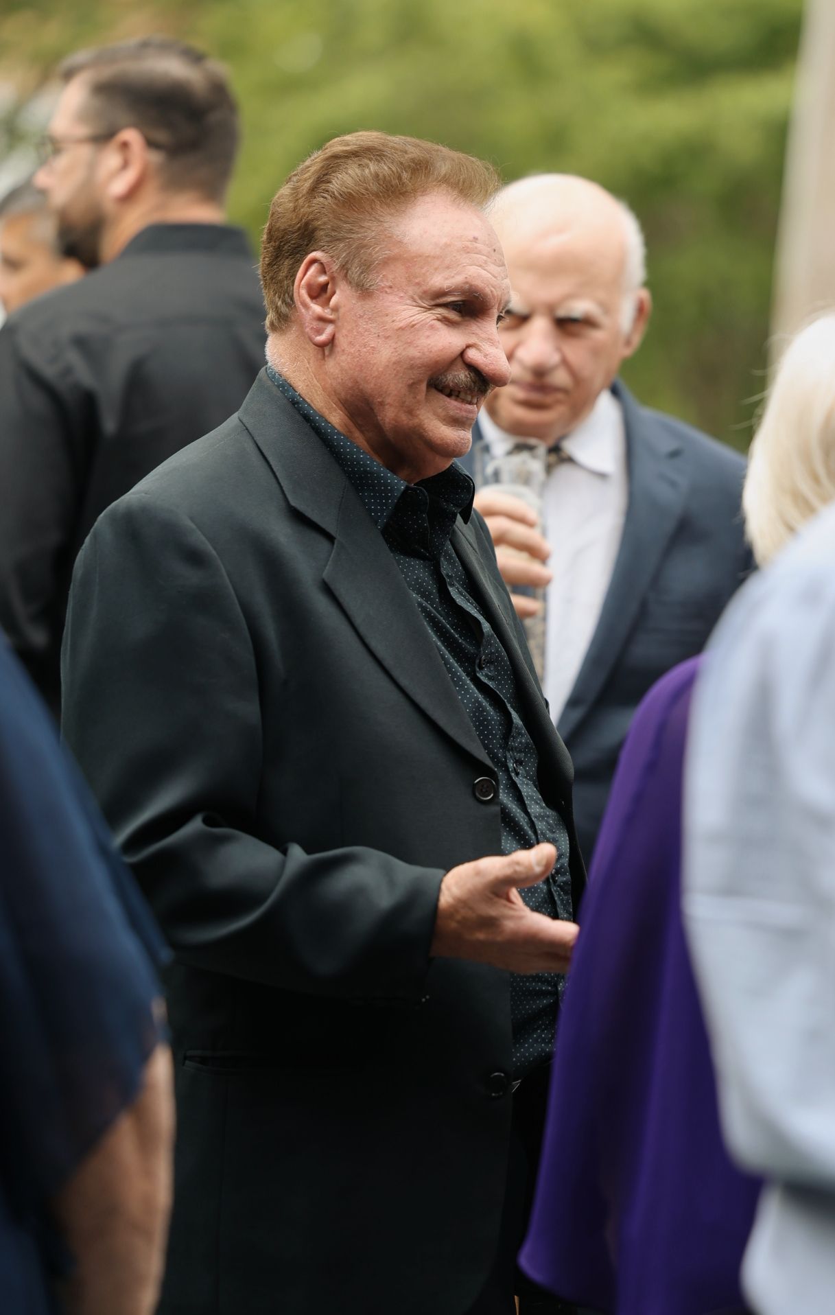 Elderly man in black suit smiling warmly while socializing at an outdoor wedding event.