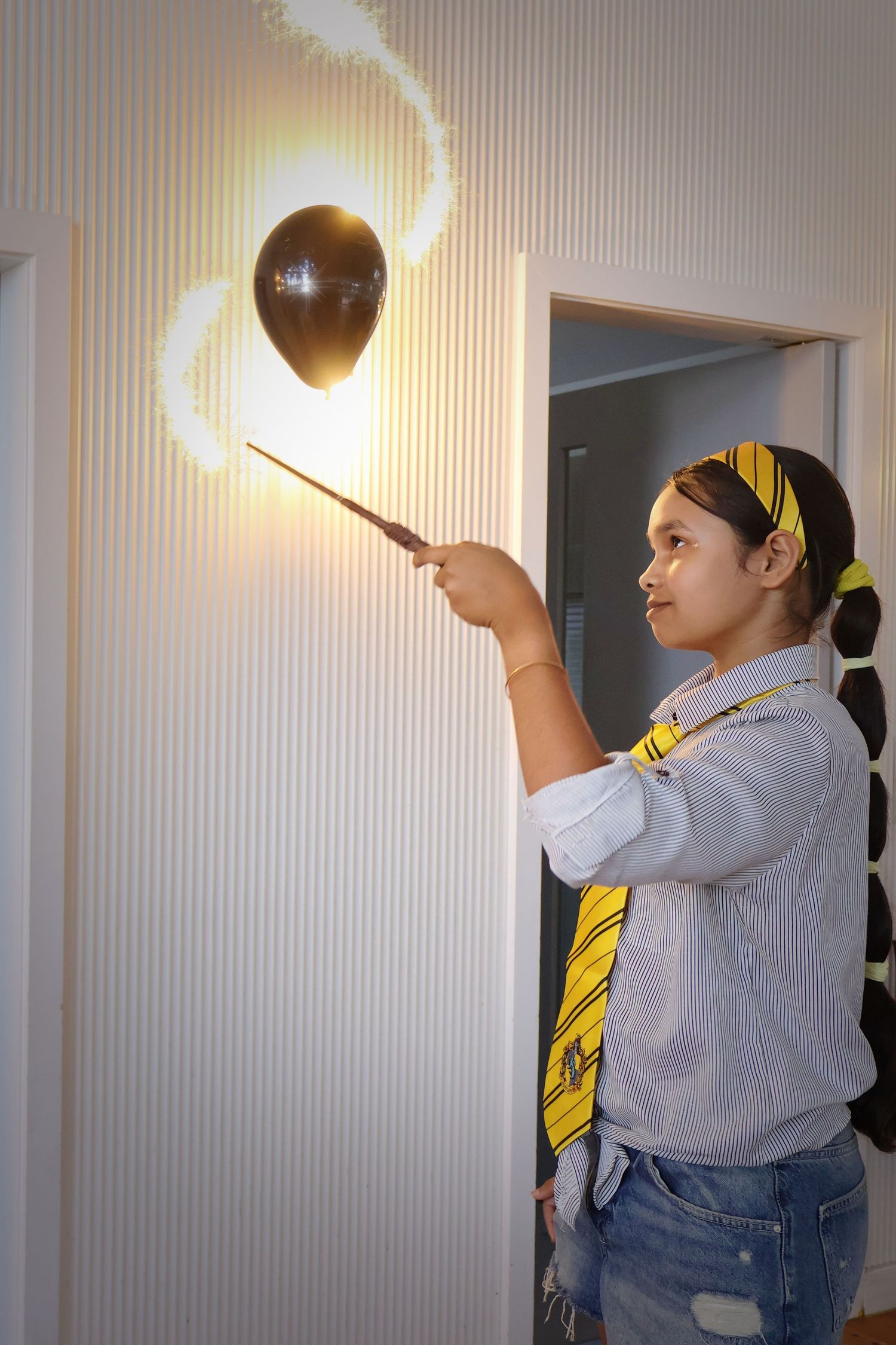 A girl in Hufflepuff tie points a magic wand at a glowing balloon on a wall.