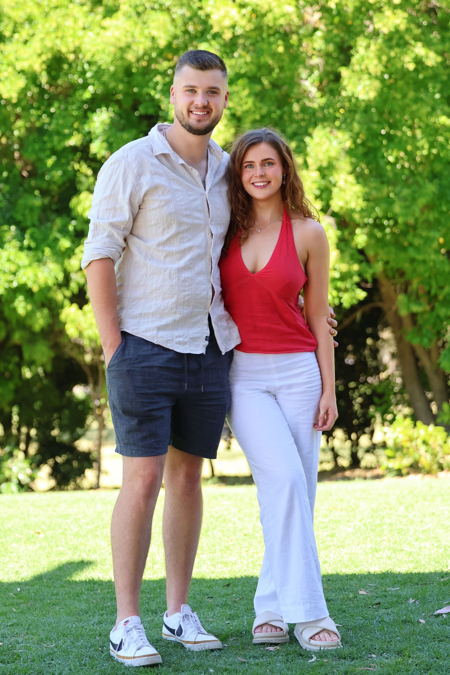 Smiling young couple posing together on green grass with lush trees, man in beige shirt and woman in red top.