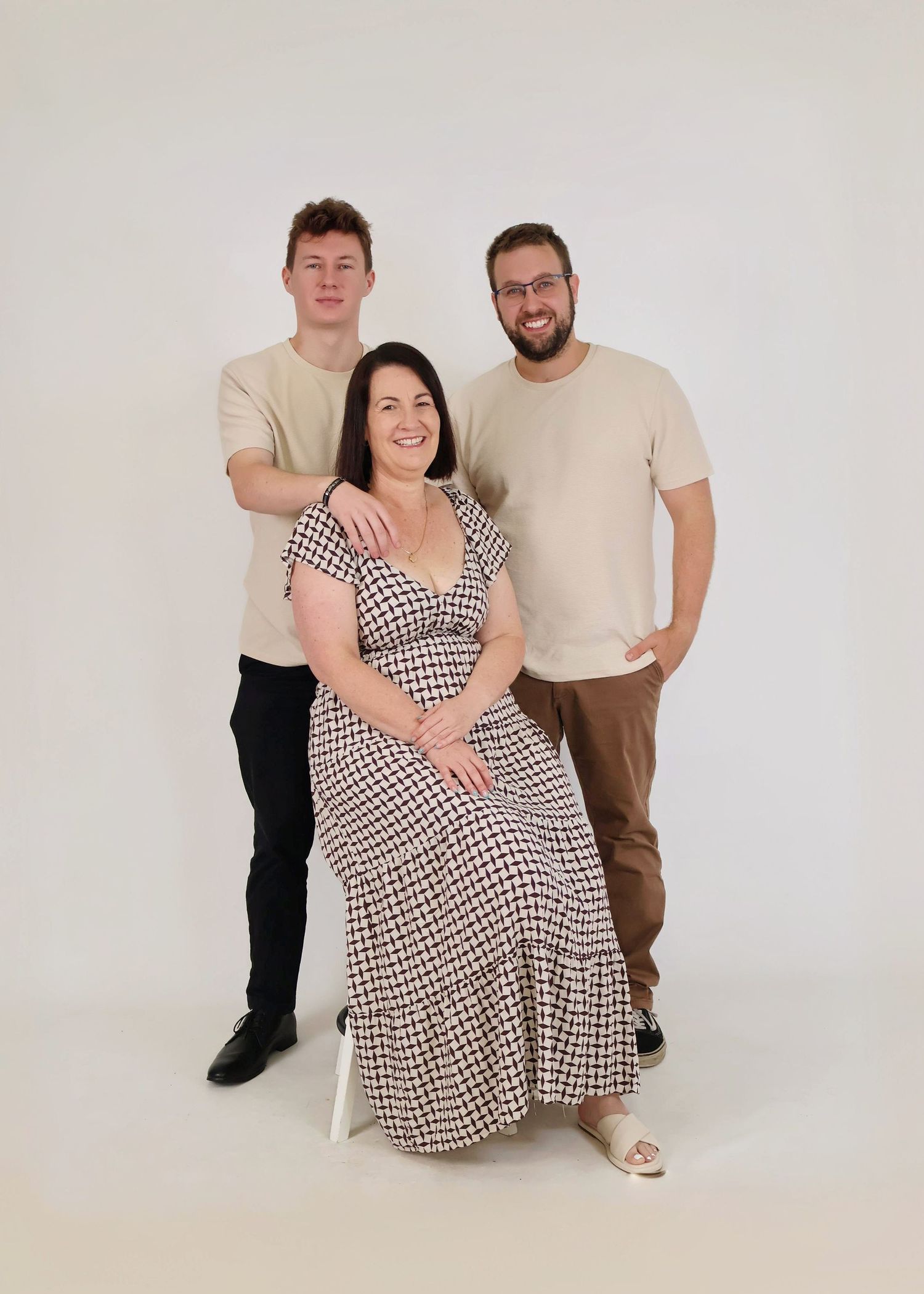 Family portrait of mother seated in patterned dress with two adult sons in a white studio.