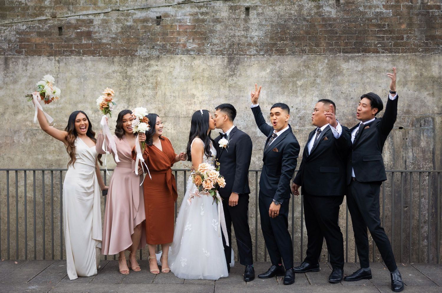 Wedding party celebrating with bouquets and cheers against rustic brick wall backdrop.
