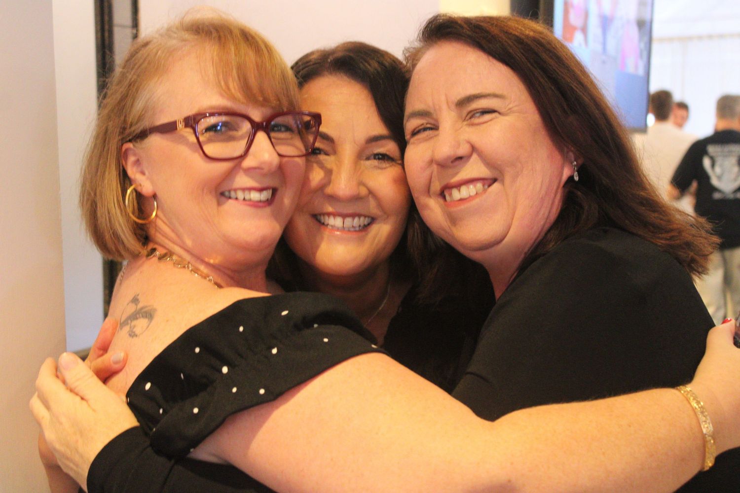 Three smiling women hugging together closely at a social gathering event.