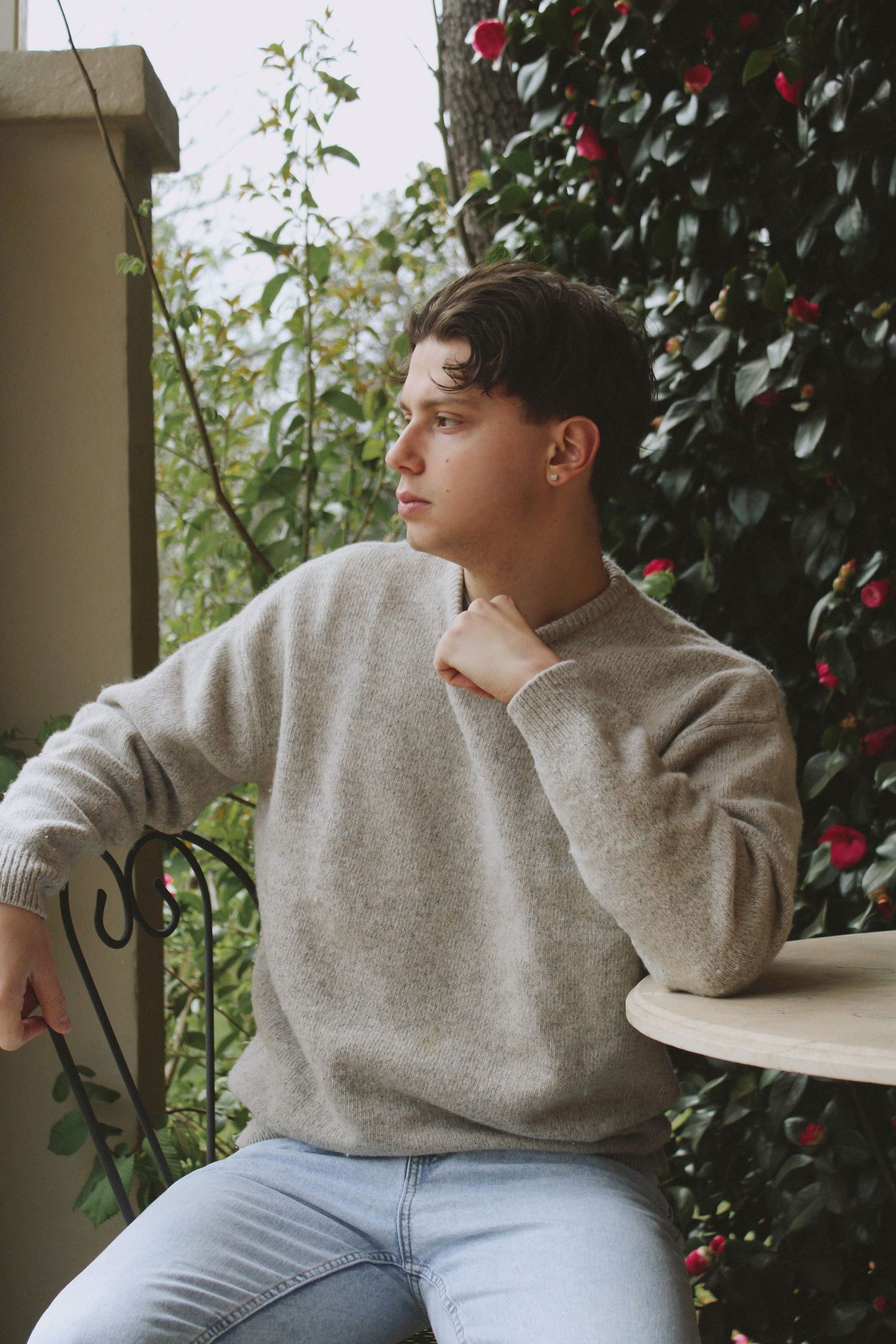 Young man in beige sweater sitting on balcony surrounded by lush green foliage and red flowers.
