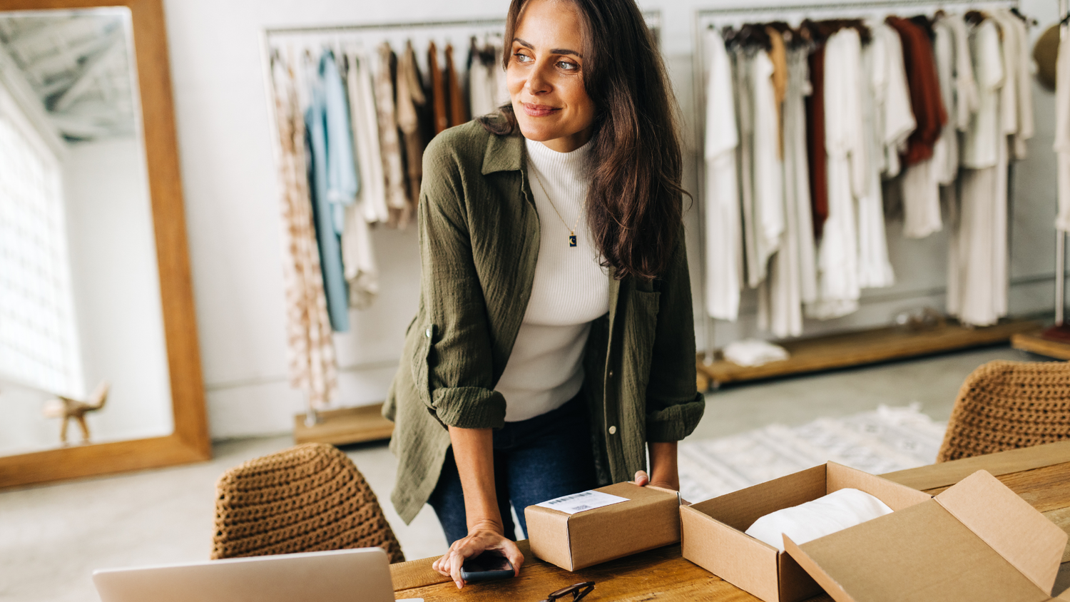 A woman in a green jacket leans over a wooden table with boxes and a laptop in a boutique clothing store.