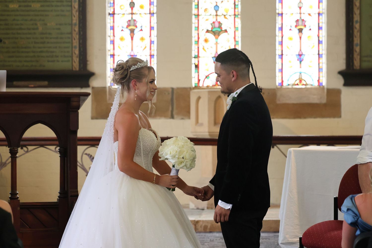 Bride and groom holding hands at church altar with colorful stained glass windows behind them.