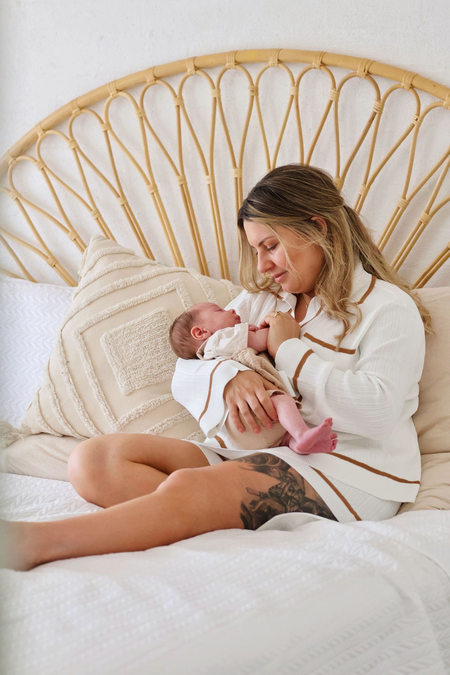 Mother in a white robe cradling her newborn baby while sitting on a bed with rattan headboard.