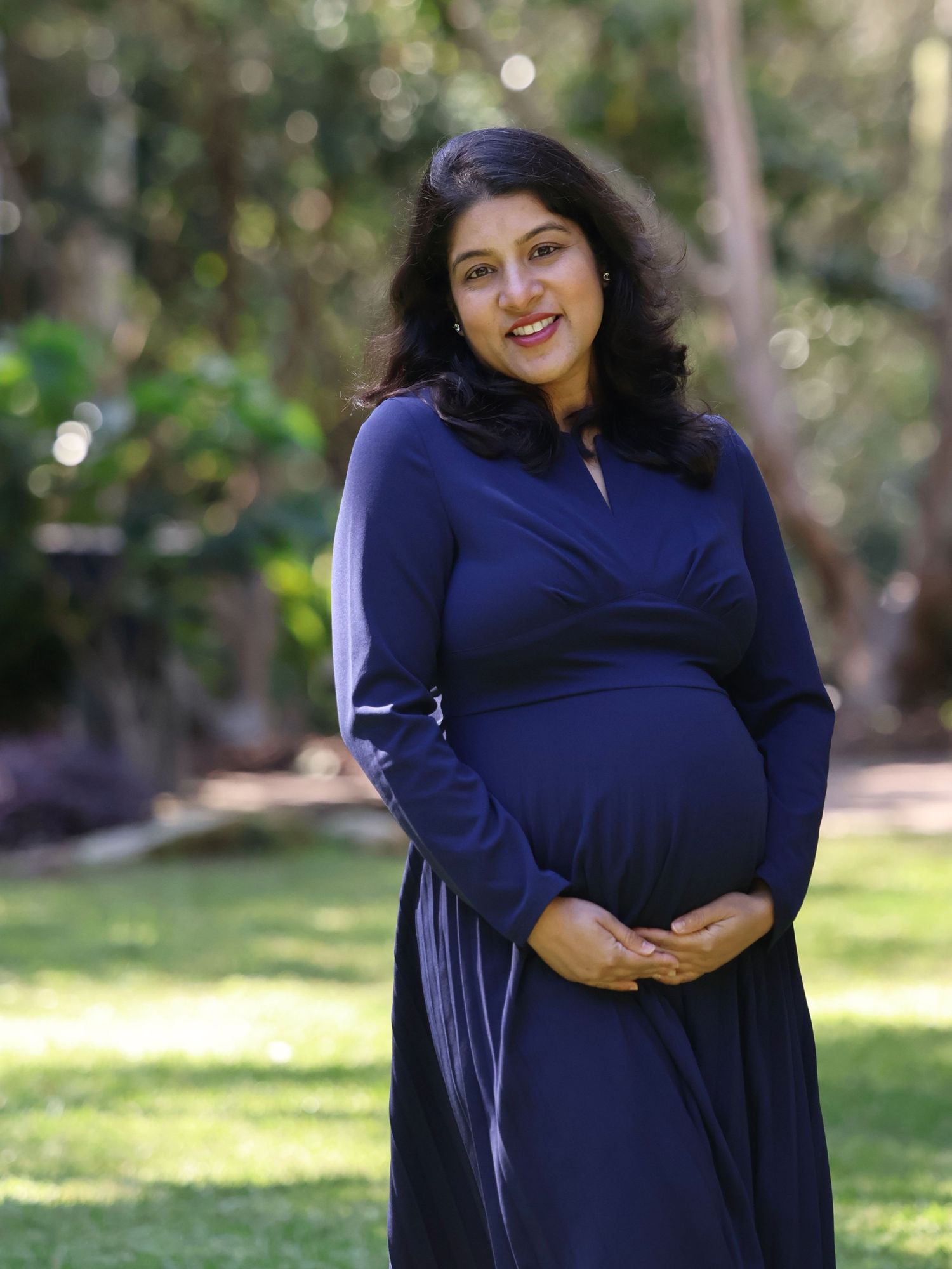 Pregnant woman in navy blue dress cradling baby bump outdoors in a lush green park setting.