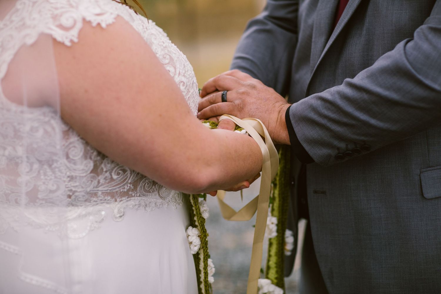 Groom tying ribbon around bride's wrist during intimate outdoor wedding handfasting ceremony.