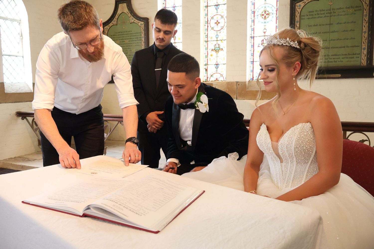 Bride and groom signing wedding register with officiant and witness in church setting.