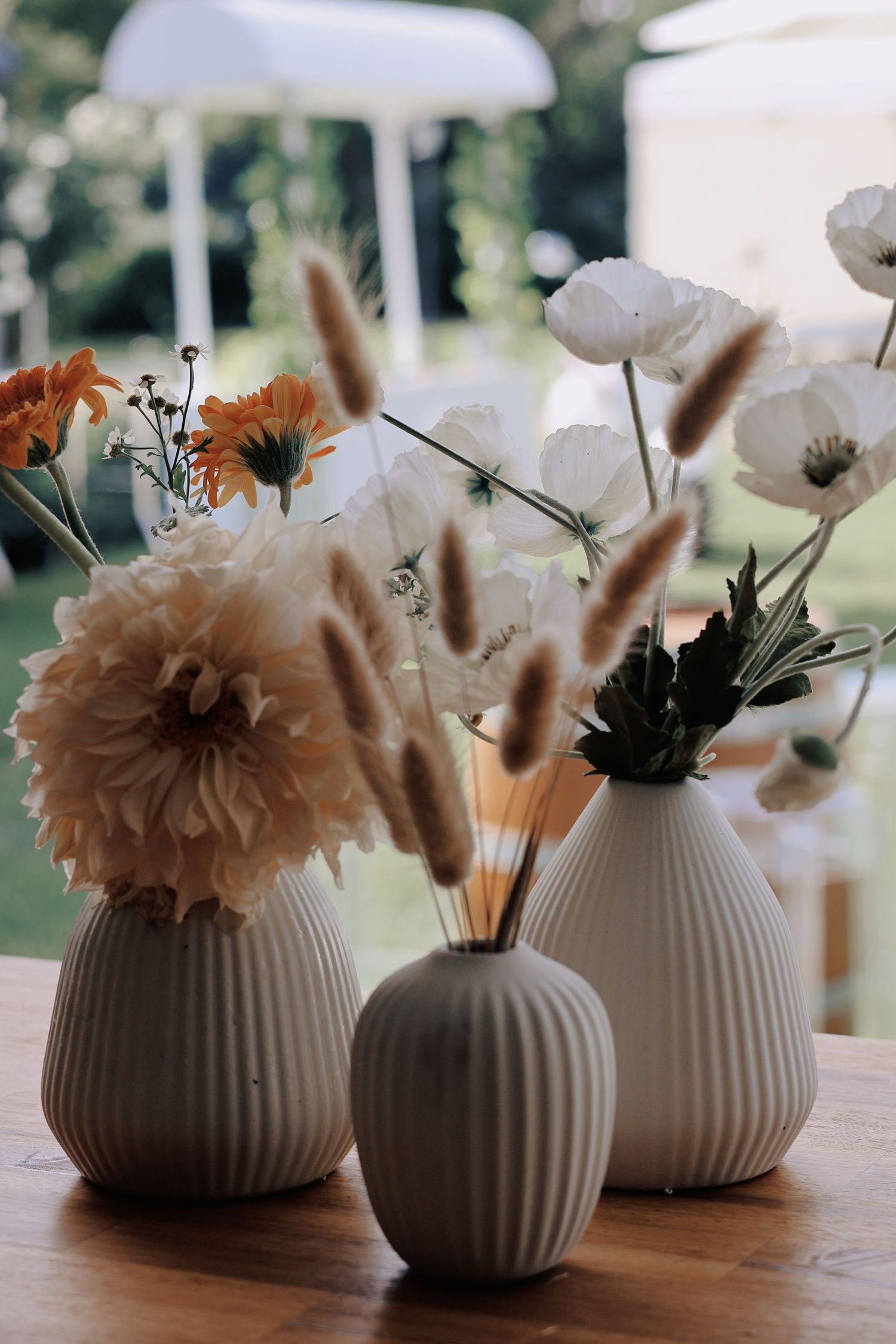 Three white ribbed ceramic vases with orange dahlias, poppies, and pampas grass on wooden table.