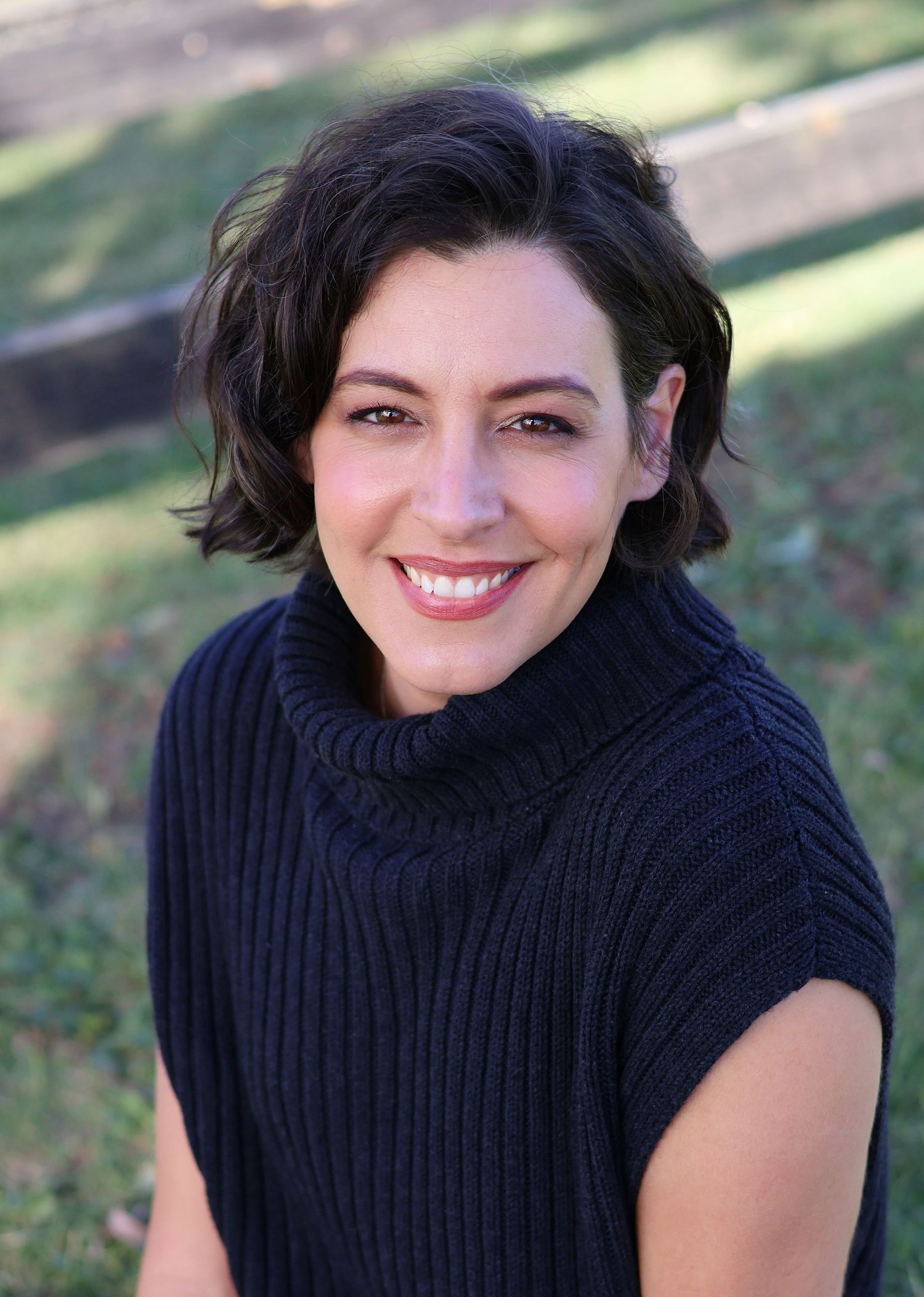 Smiling woman with short dark hair wearing a navy turtleneck sweater vest in an outdoor portrait.