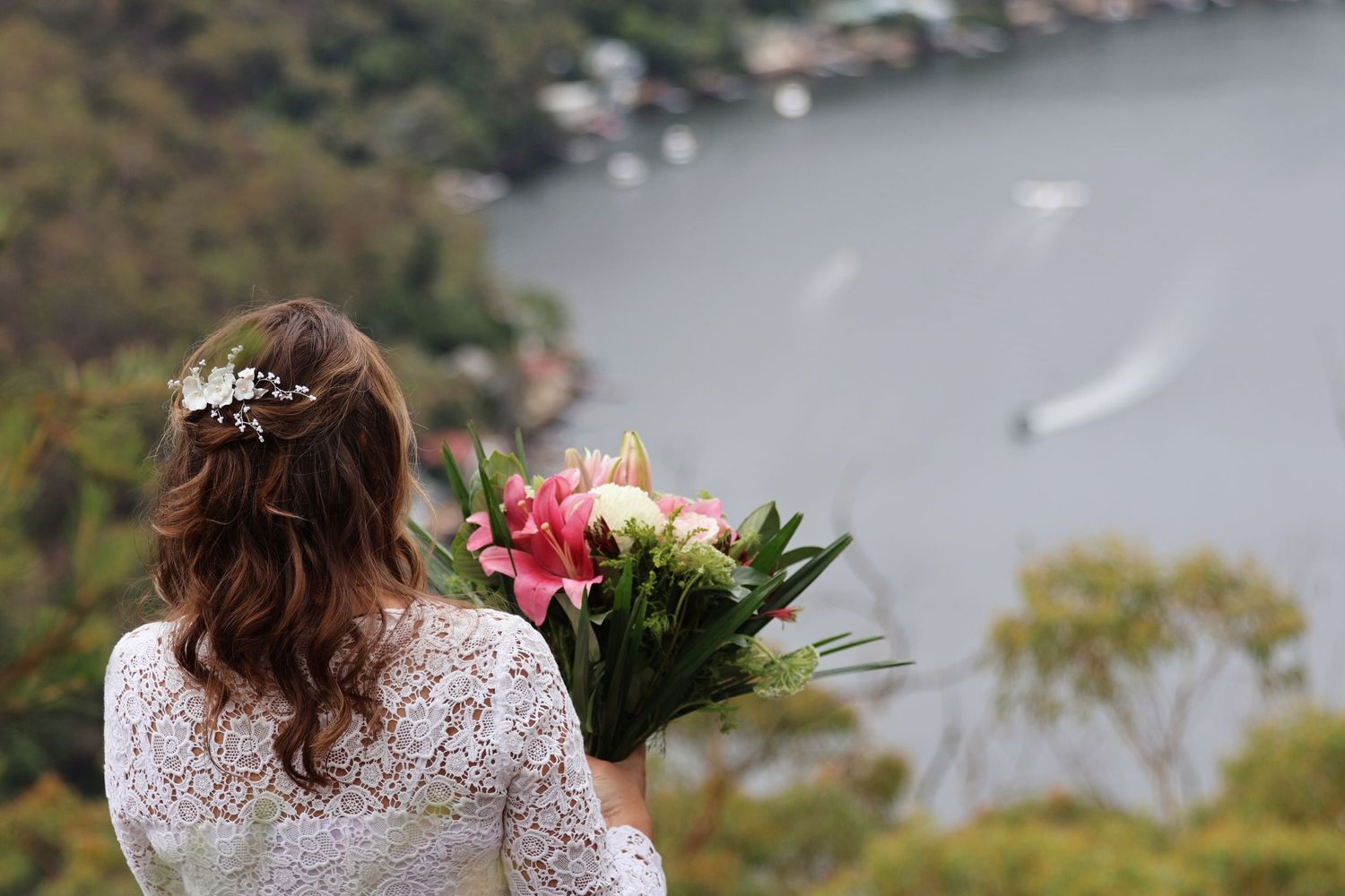 Bride in white lace dress holding pink lily bouquet overlooking a scenic harbor from a hilltop viewpoint.