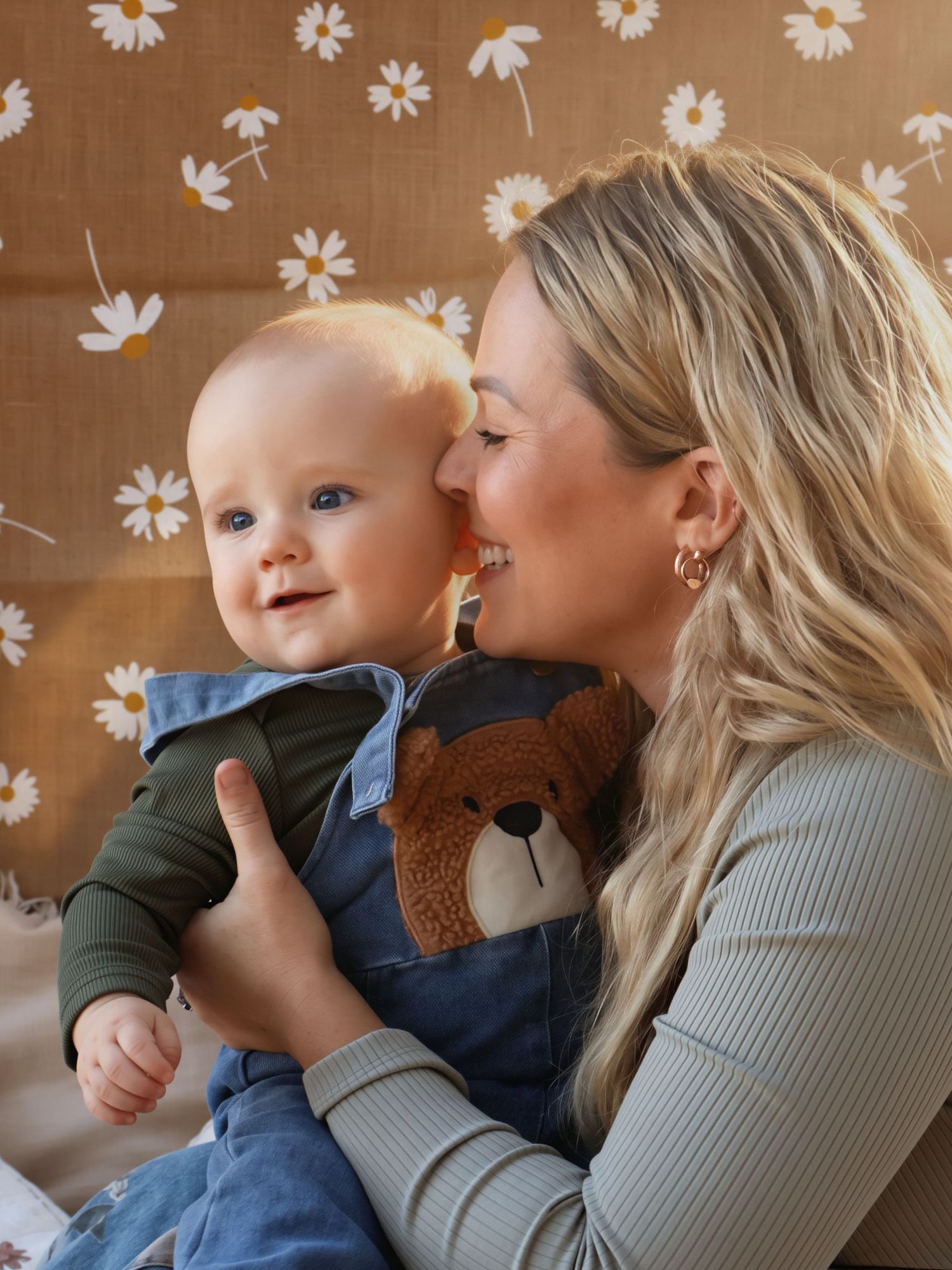Blonde mother kissing smiling blue-eyed baby holding teddy bear against daisy wallpaper.
