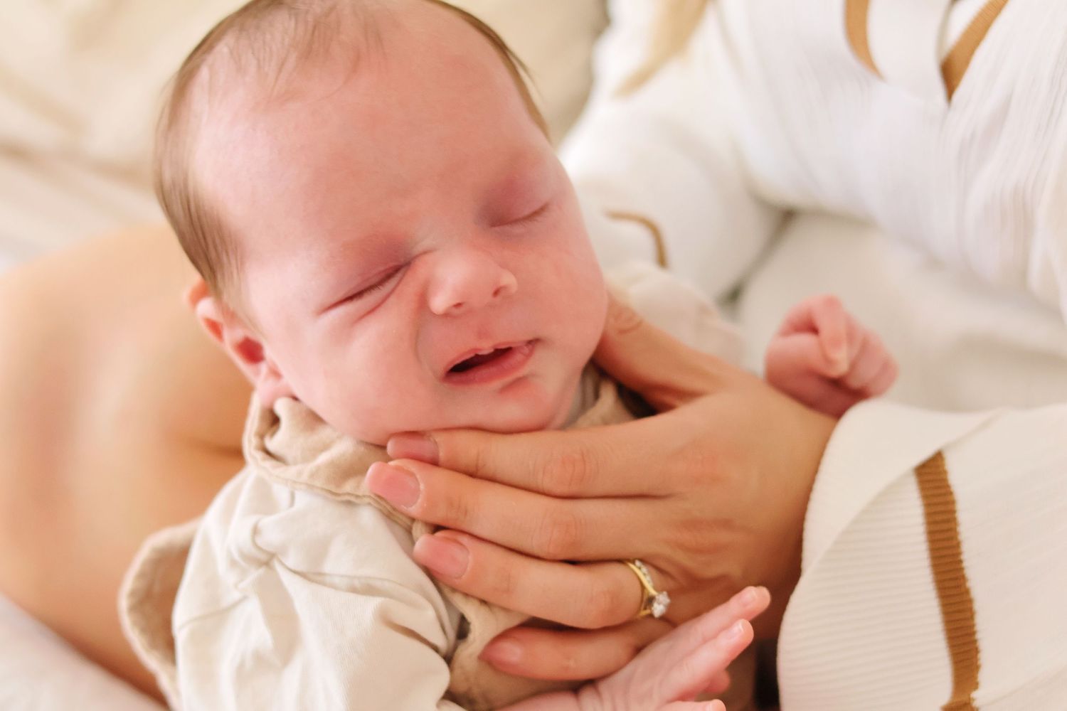 Crying newborn baby in cream outfit gently held in mother's hands.
