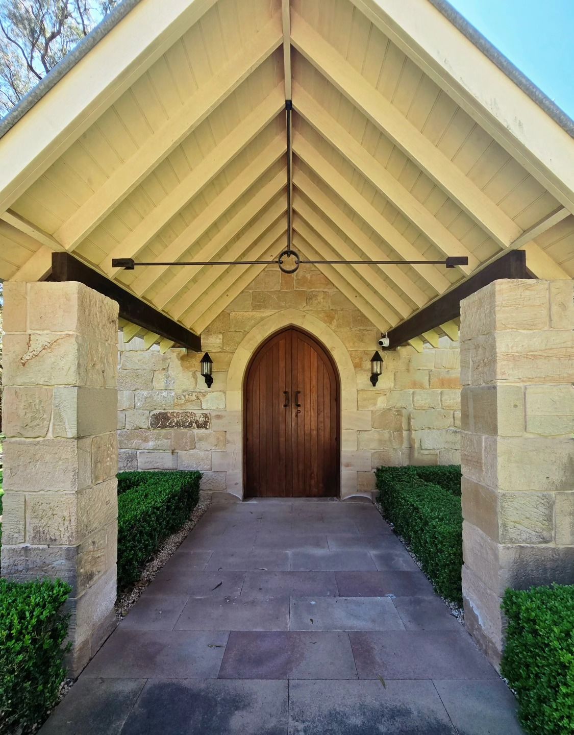 Elegant stone chapel entrance with arched wooden doors, lanterns, and manicured hedges under a gabled roof.