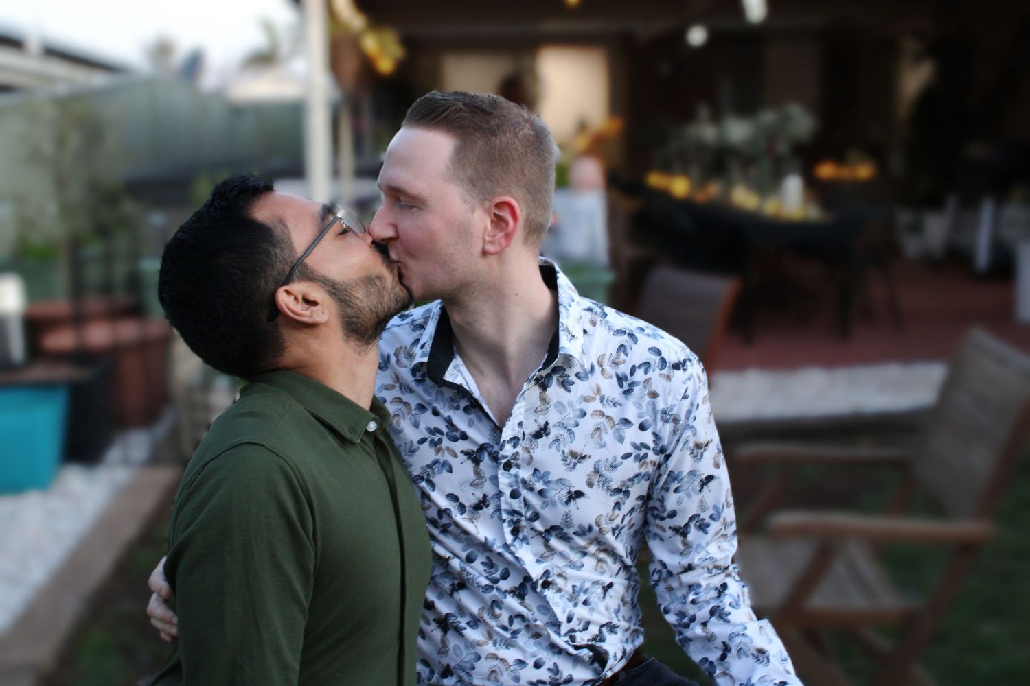 Two men sharing a tender kiss outdoors in a garden setting with warm evening lights in the background.