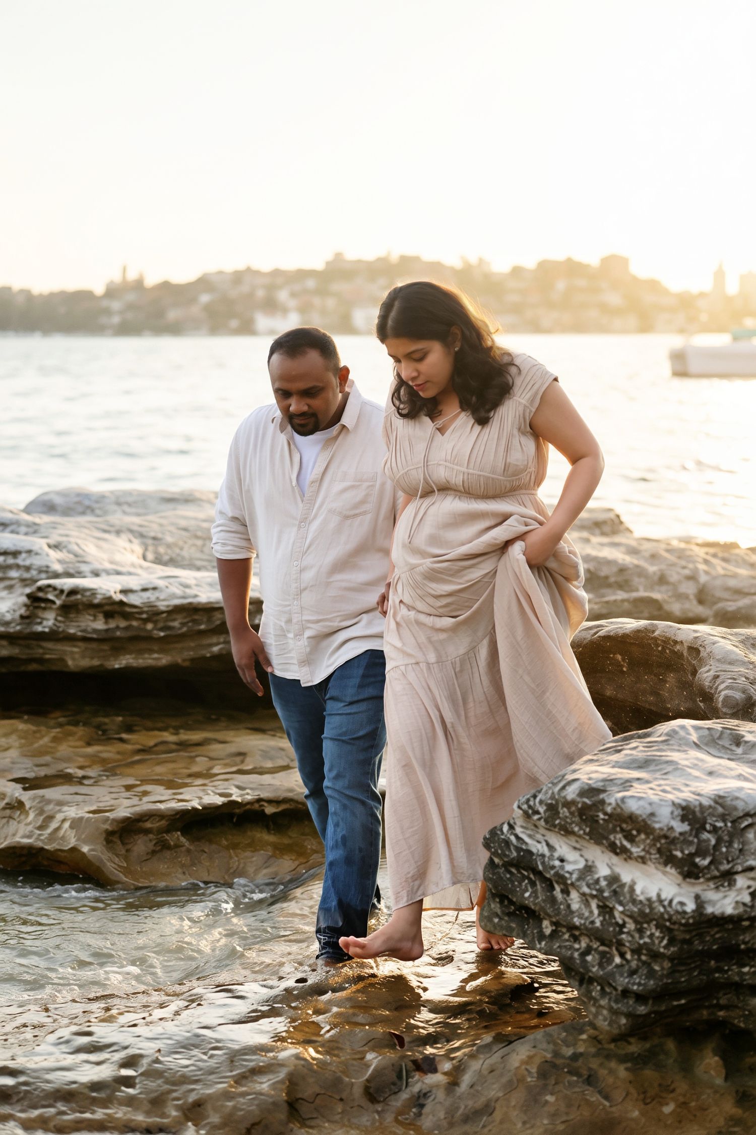 Pregnant couple walks carefully over coastal rocks at golden sunset near a scenic waterfront.