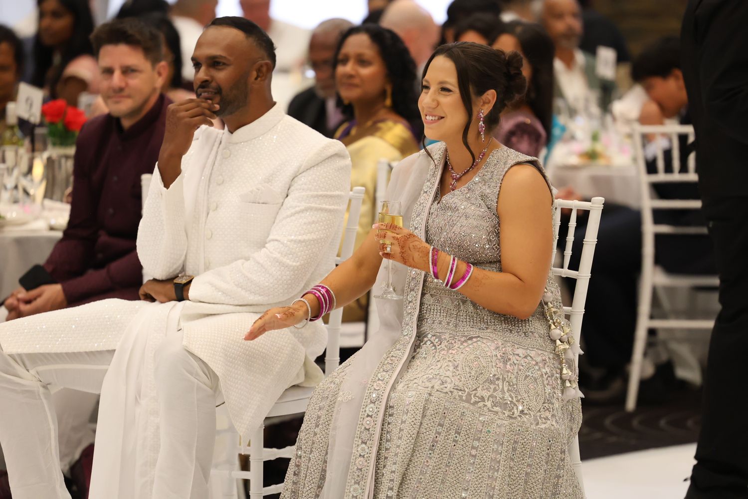 Bride and groom in white and silver Indian wedding attire sit together during elegant reception celebration.