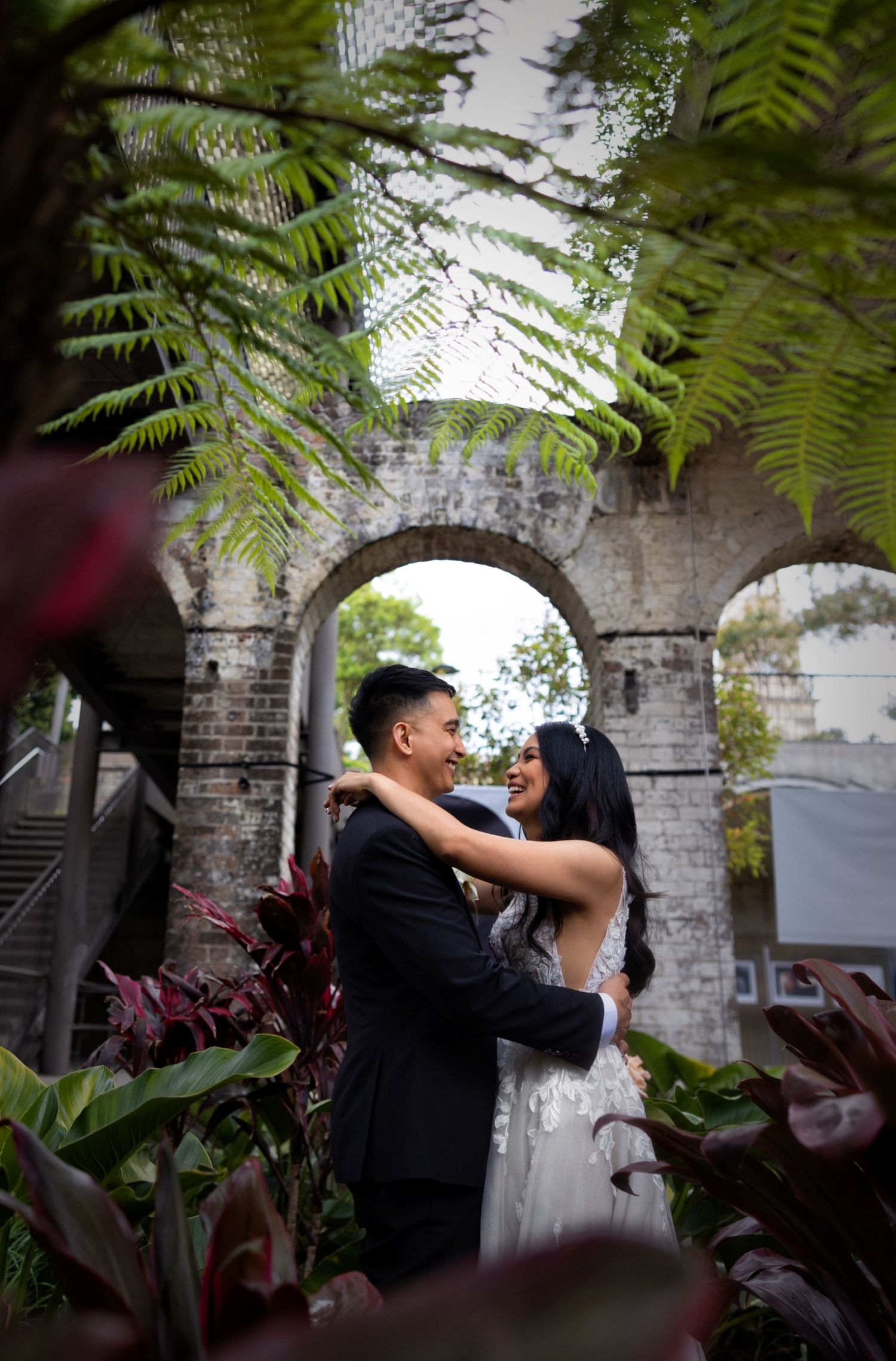 Romantic couple embracing in garden framed by fern leaves and historic brick arch.