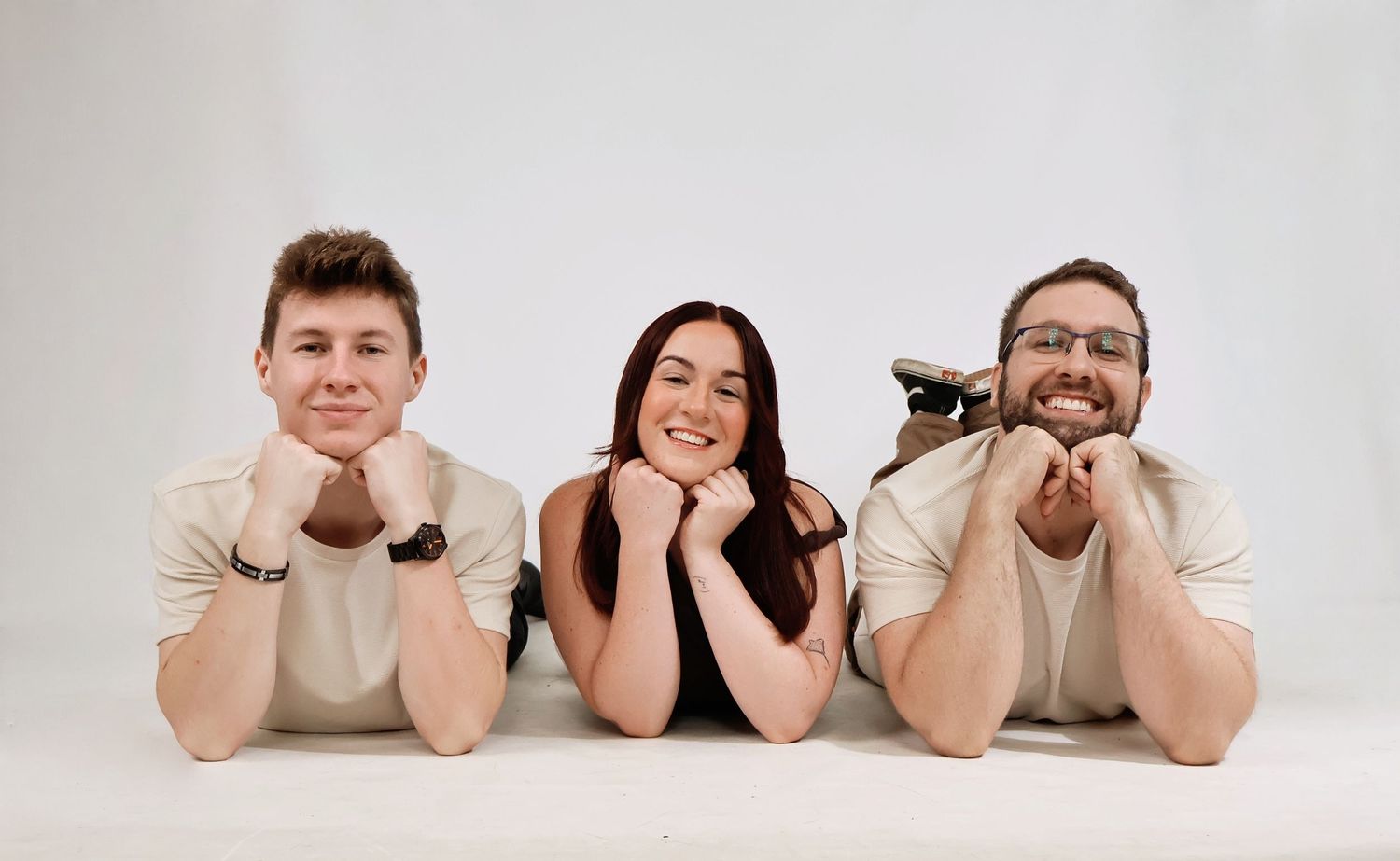 Three smiling siblings lying on floor with chins resting on hands in a bright white studio.