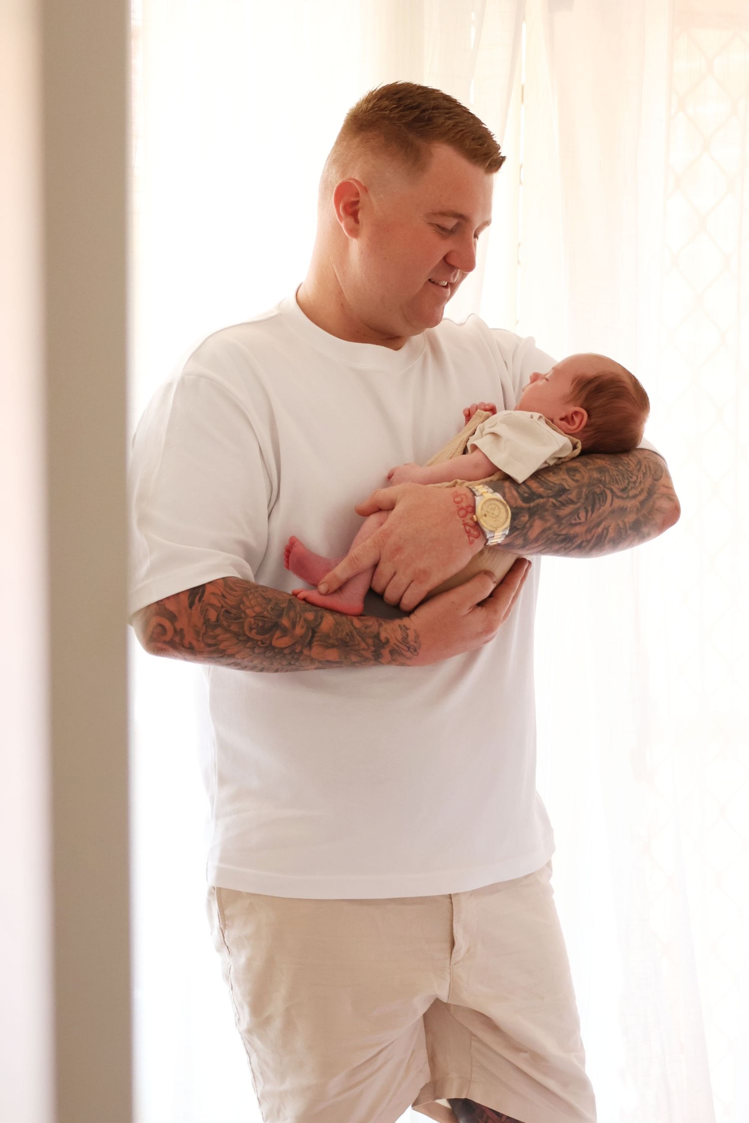 Tattooed father in white t-shirt gently cradling sleeping newborn baby by bright window.