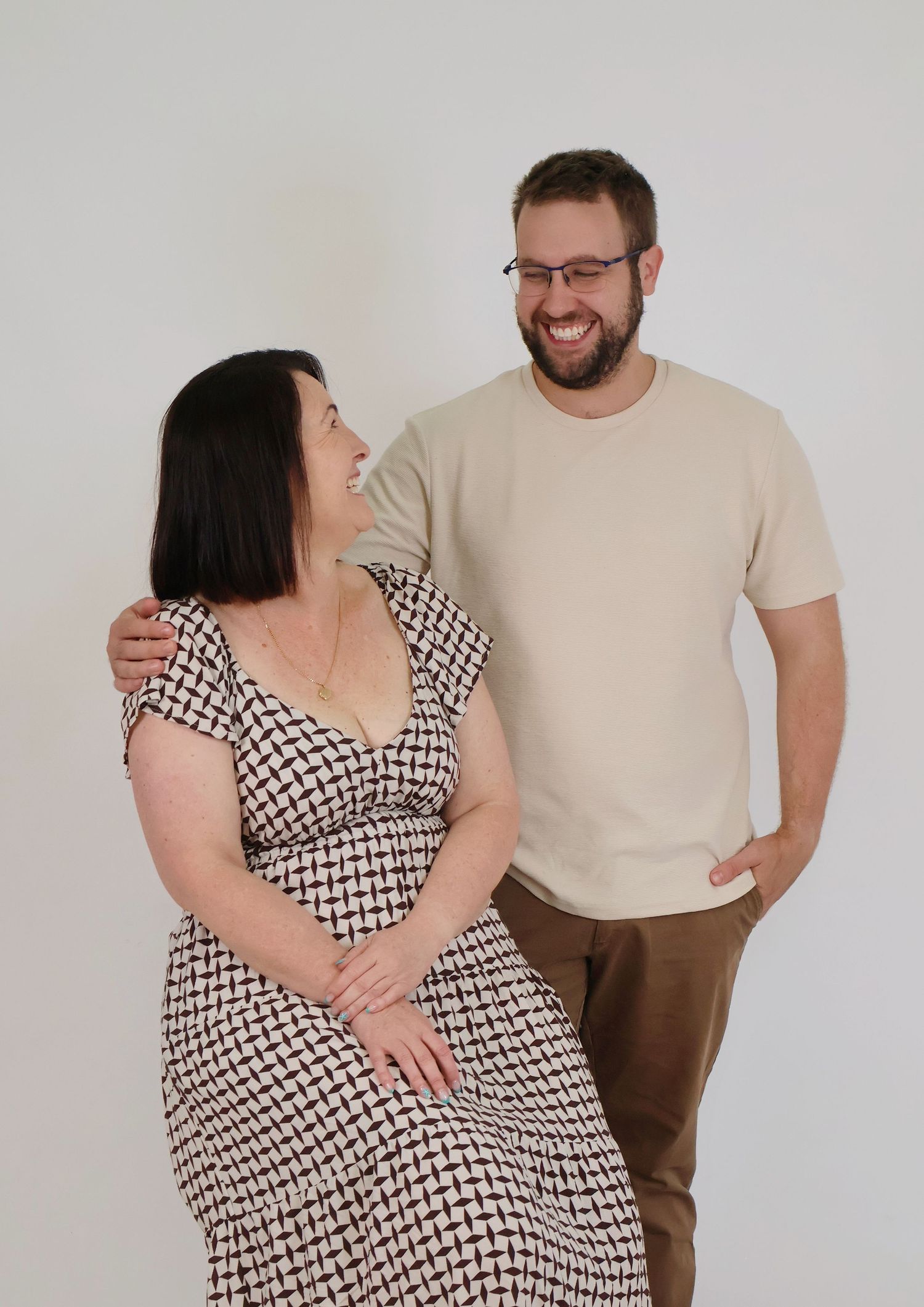 Smiling bearded man with glasses and woman in patterned dress posing together on white background.